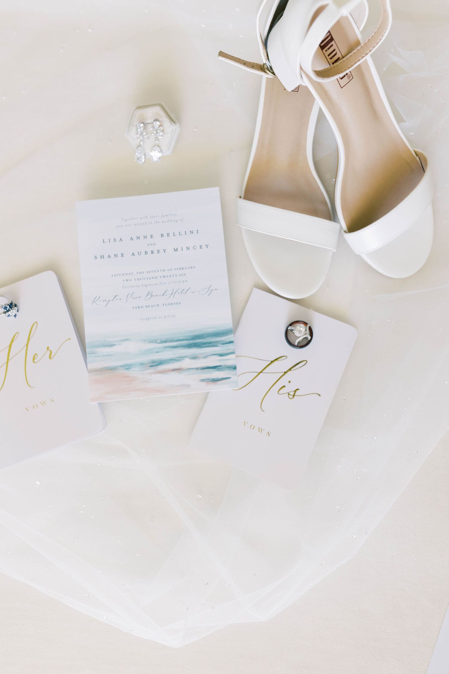 White bridal heels, wedding invitation with watercolor beach design, and His and Hers vow books arranged on white fabric.