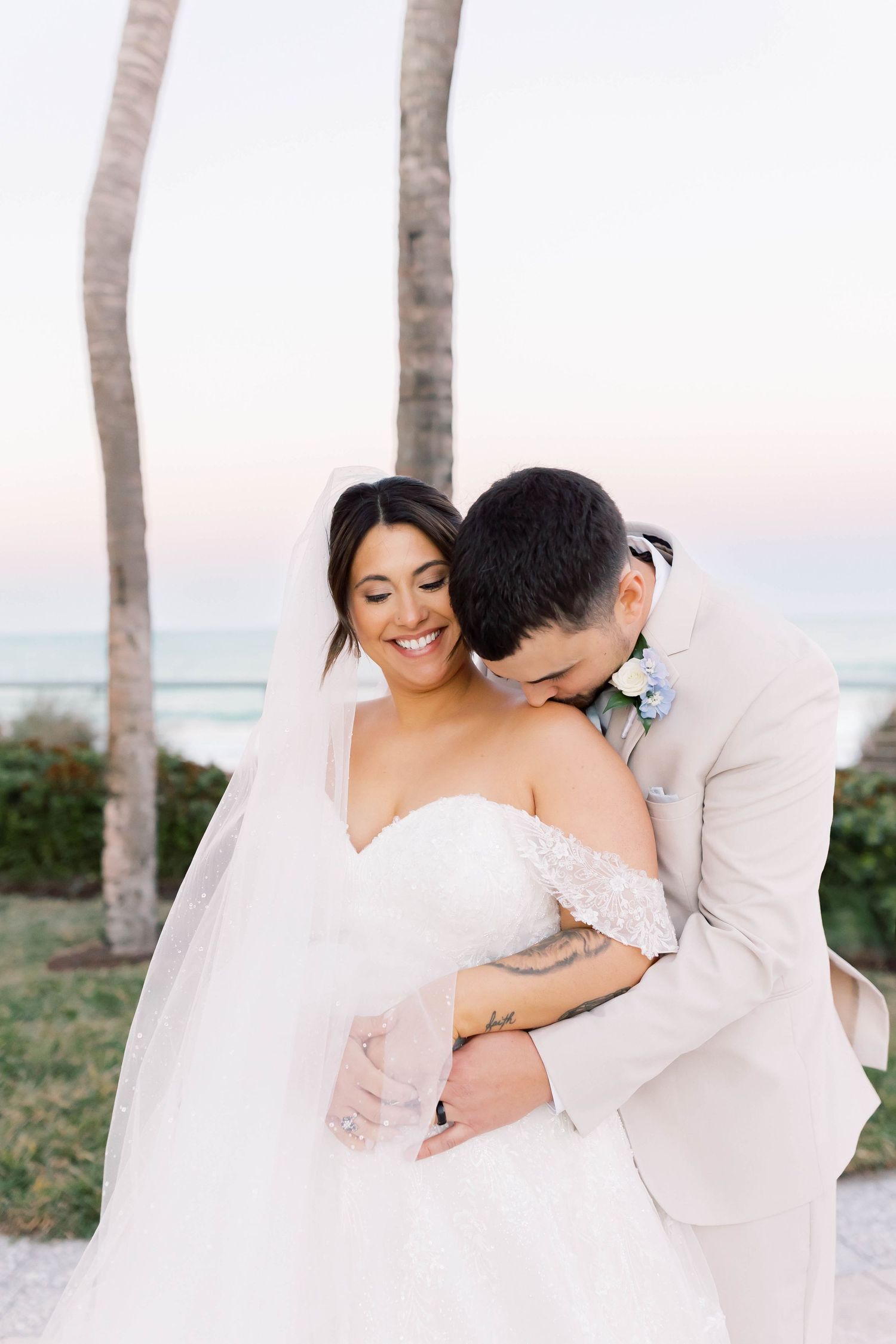 Smiling bride embraced by groom in tan suit with ocean and palm trees in background.