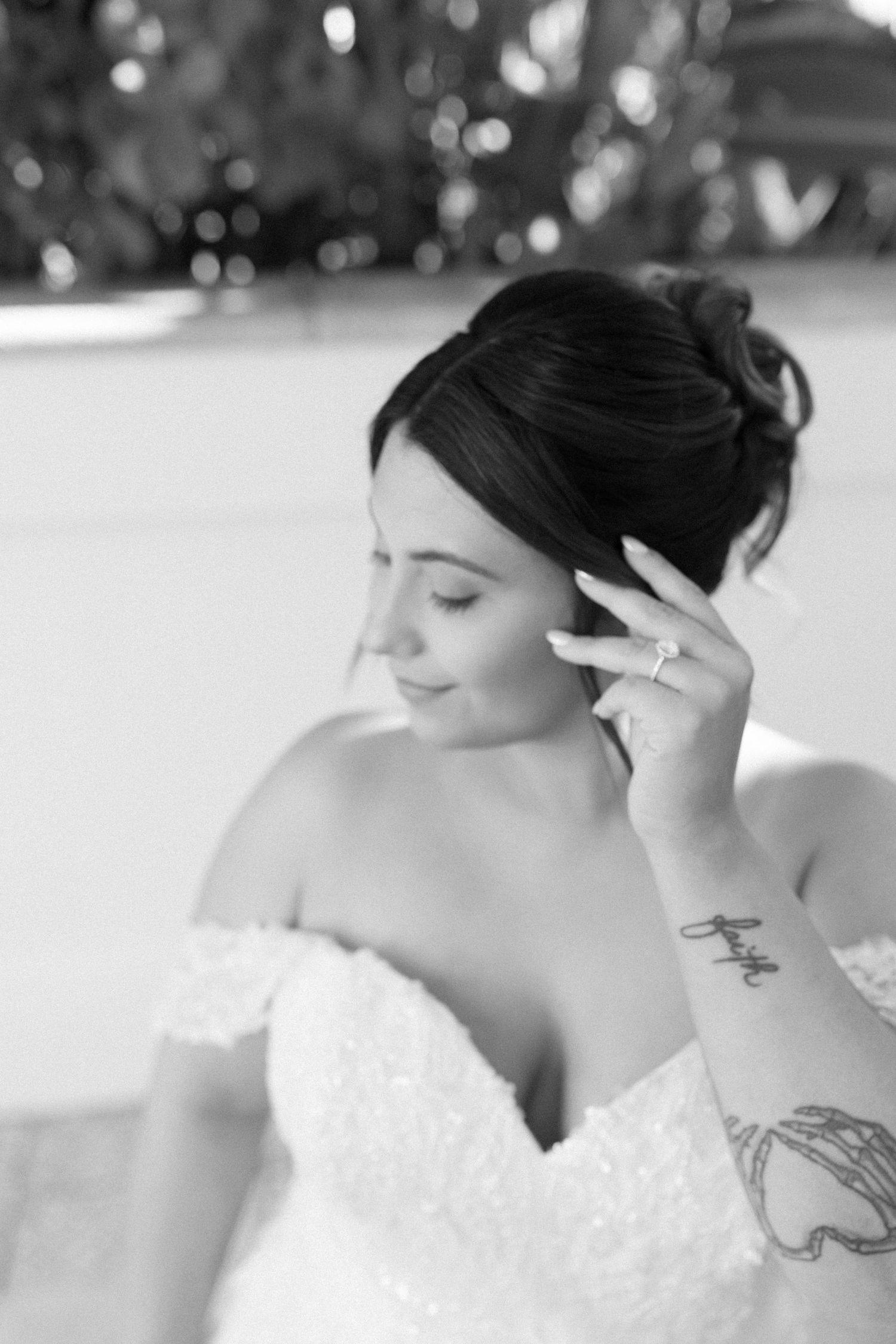 Black and white bridal portrait of a bride in off-shoulder lace gown, adjusting earring, with tattoo visible on wrist.