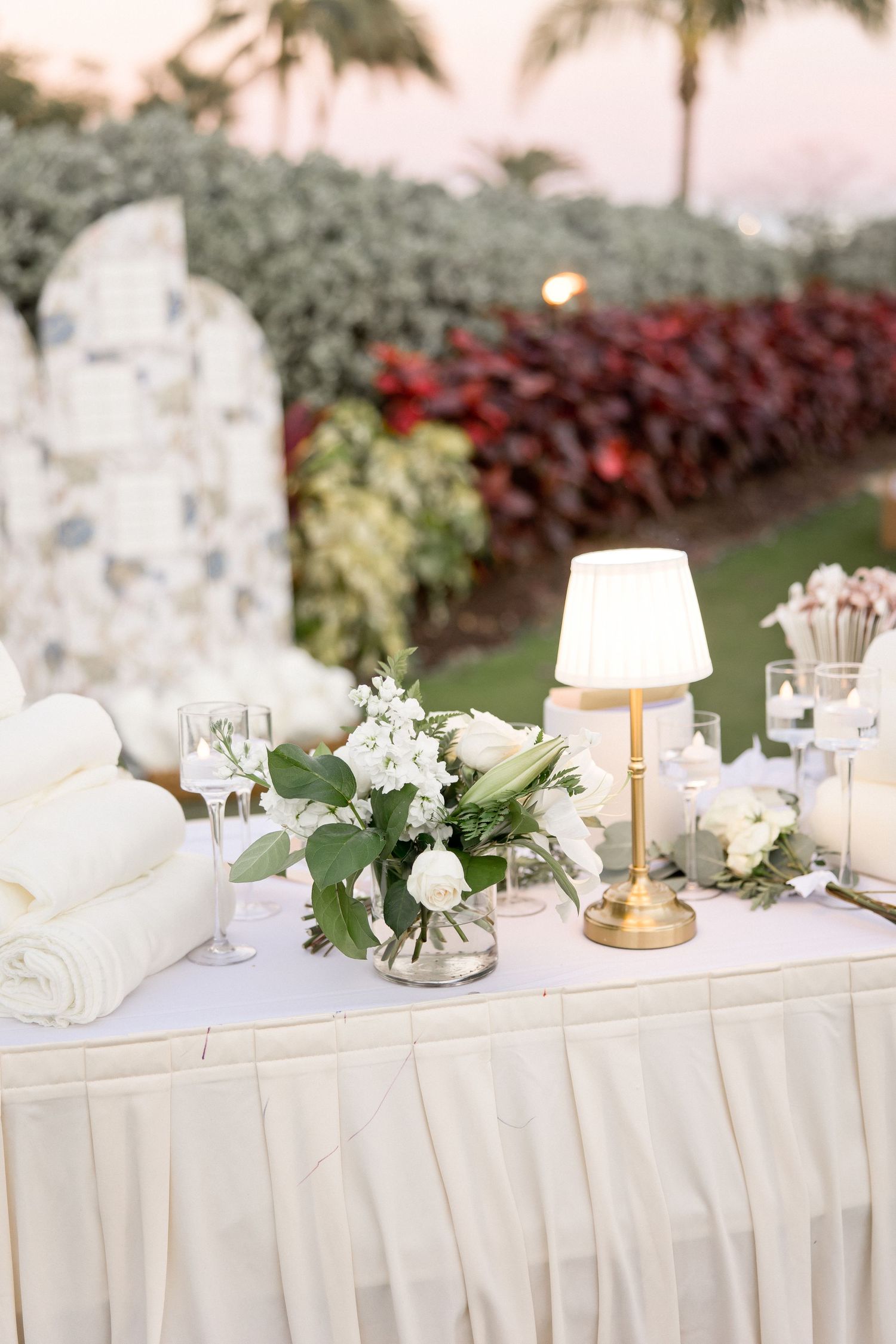Elegant white wedding reception table decorated with floral arrangements and candles against tropical landscaping.