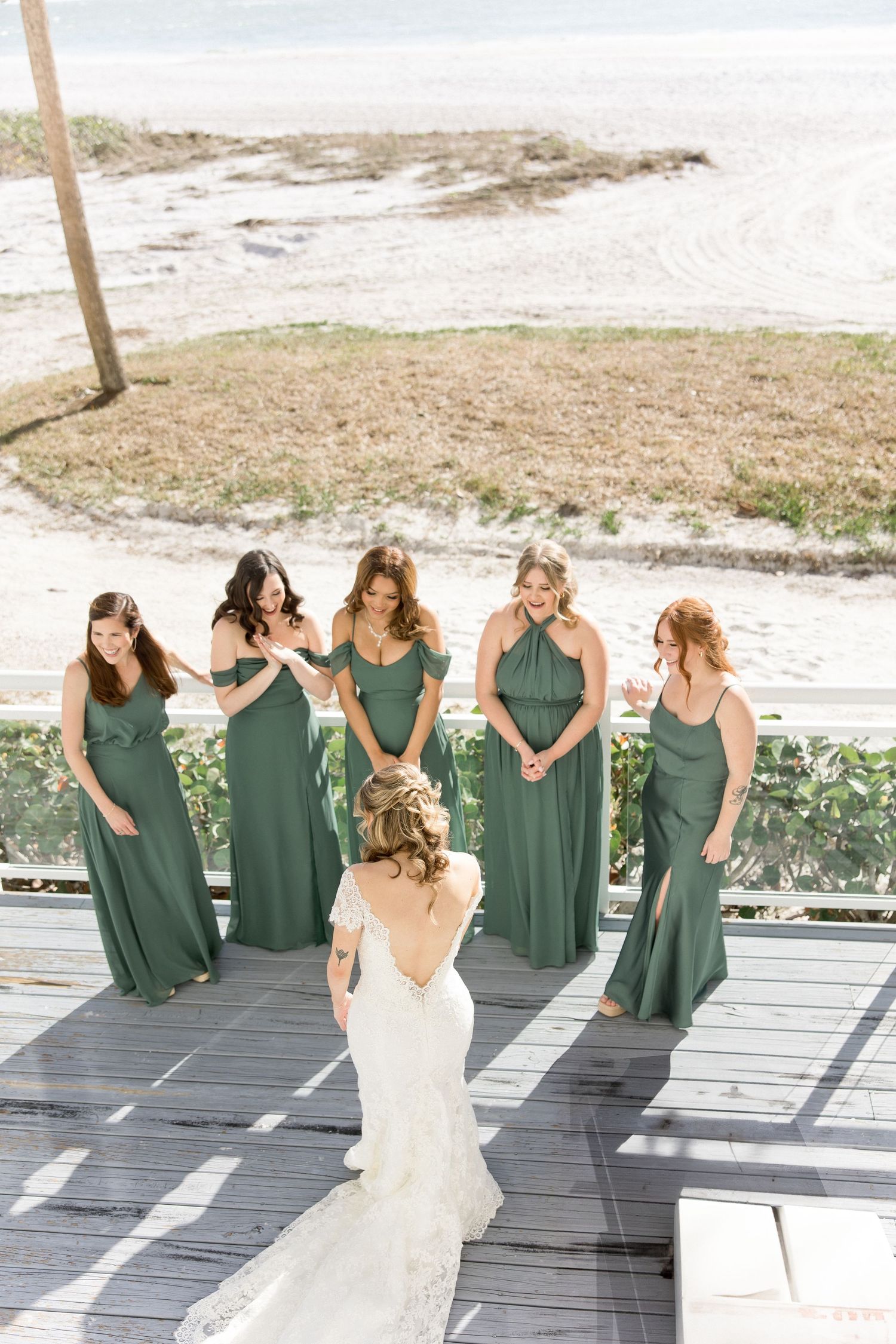 Bridesmaids in sage green dresses dance and celebrate on a wooden deck by the beach.