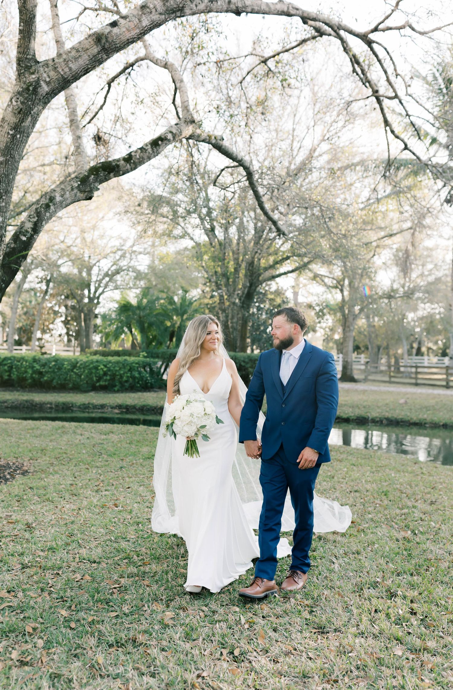Wedding couple standing by pond under winter trees in outdoor setting.