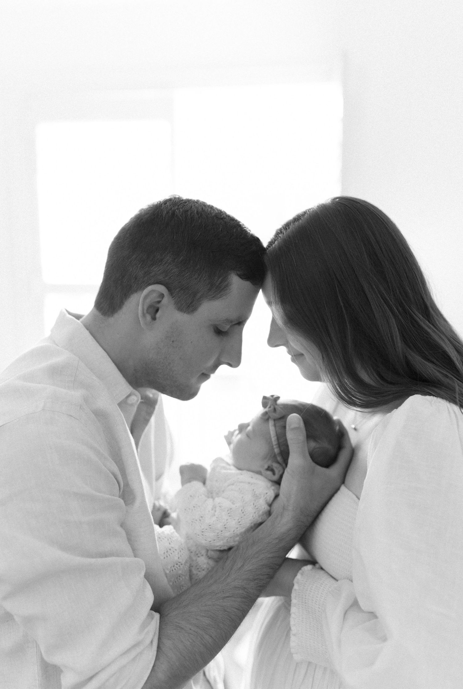 Black and white portrait of parents cradling their newborn with foreheads touching.
