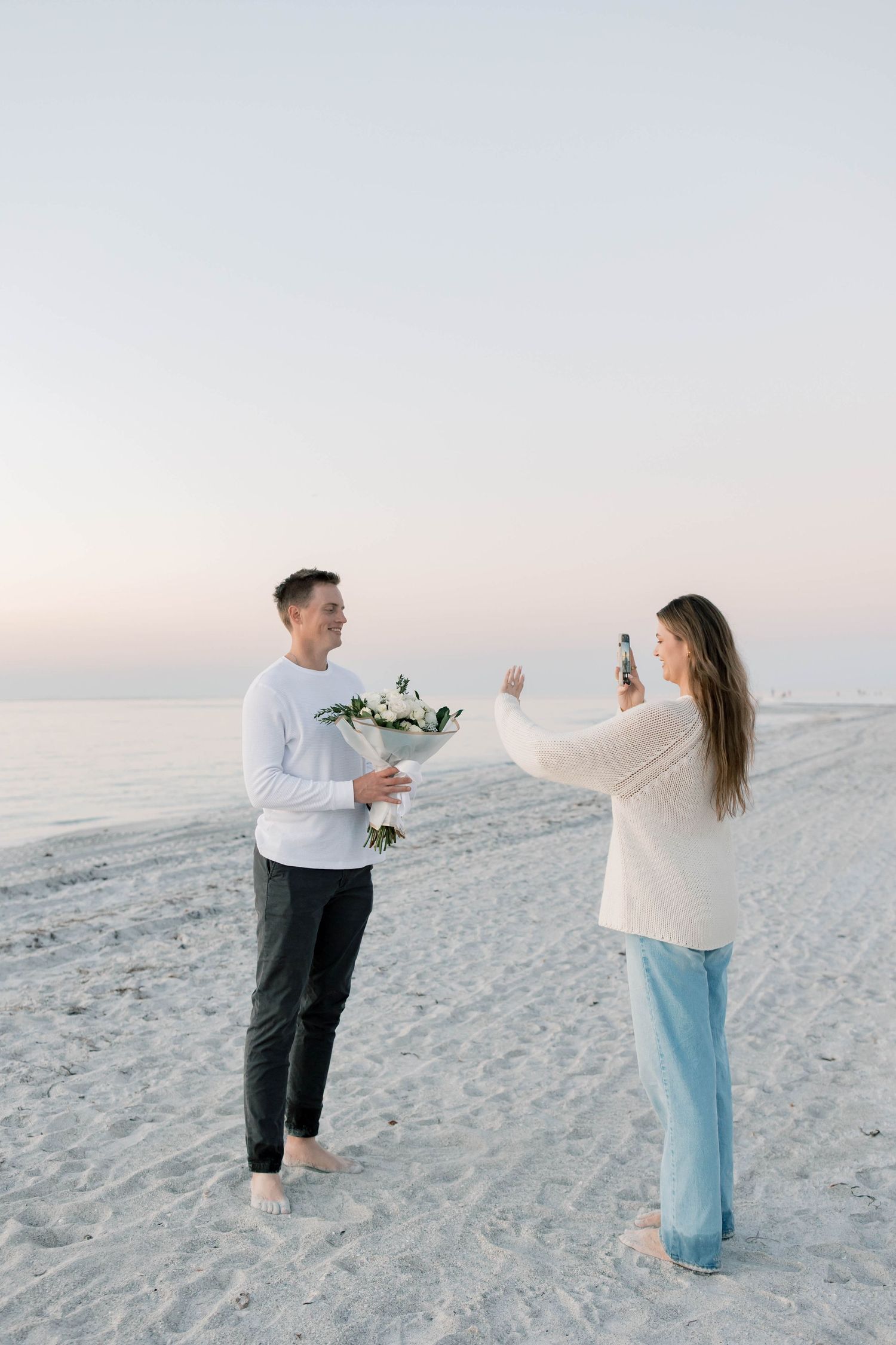 Couple celebrating with flowers during beach proposal at sunset.