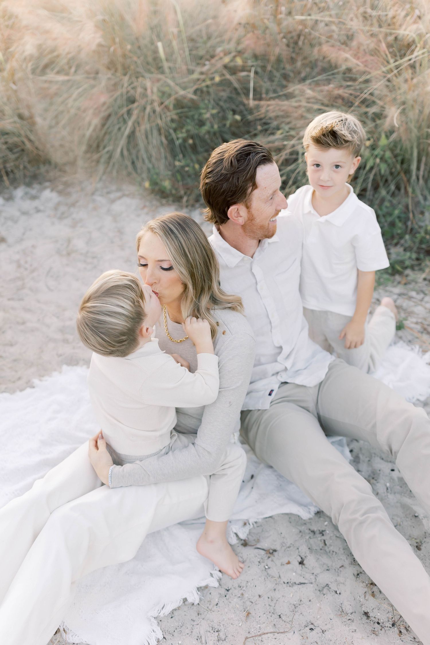 Family dressed in neutral colors shares tender moments on sandy beach during golden hour.