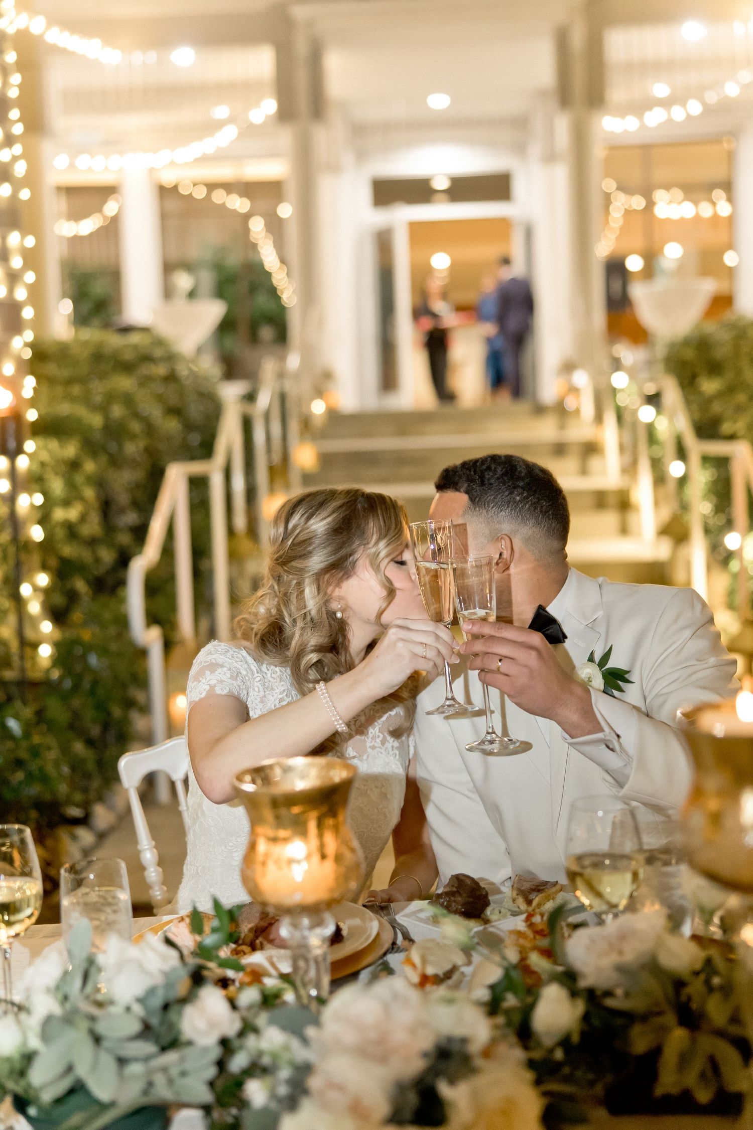 Newlyweds share an intimate champagne toast at their reception with string lights and floral decor.
