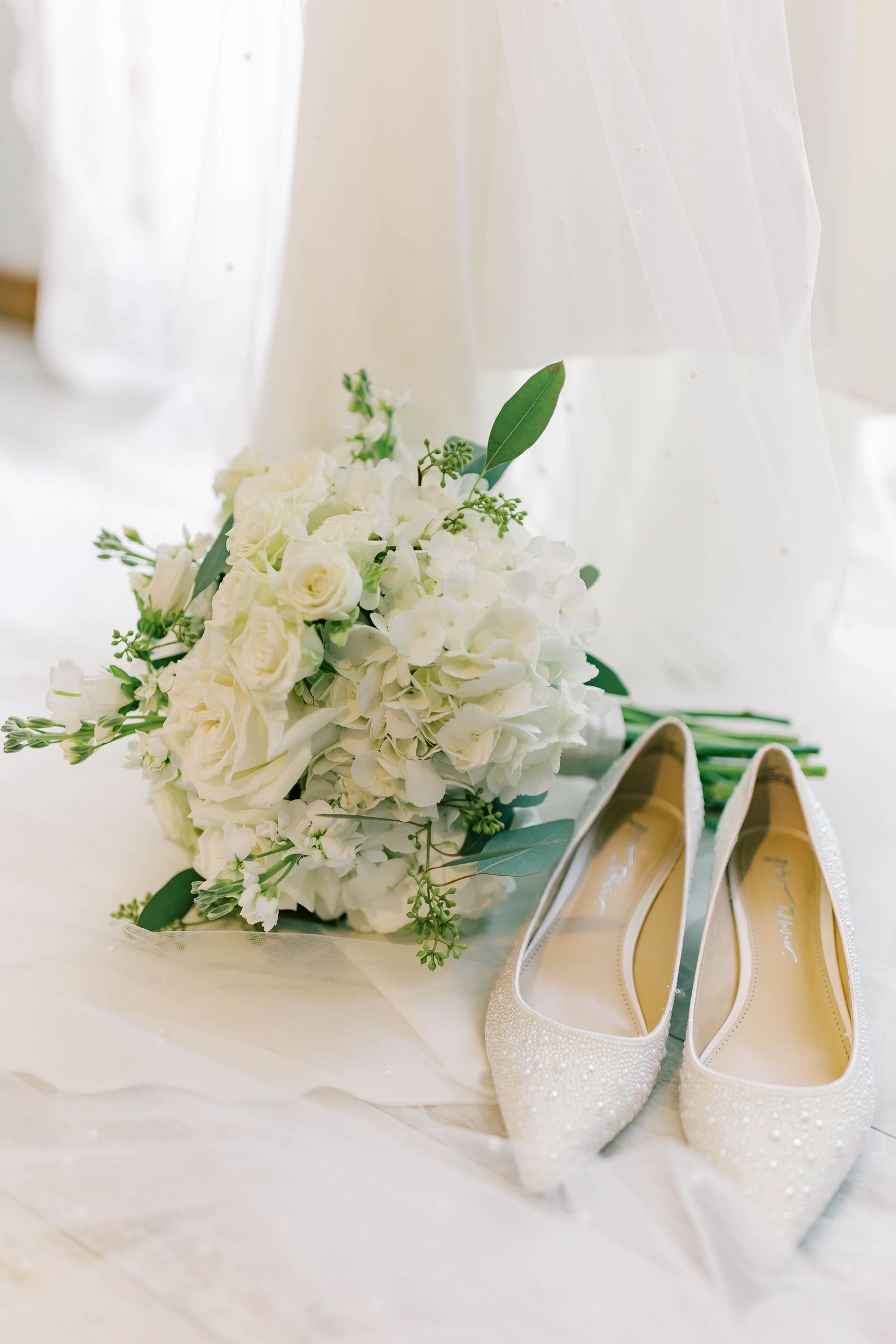 Elegant bridal shoes with white roses and greenery arrangement.