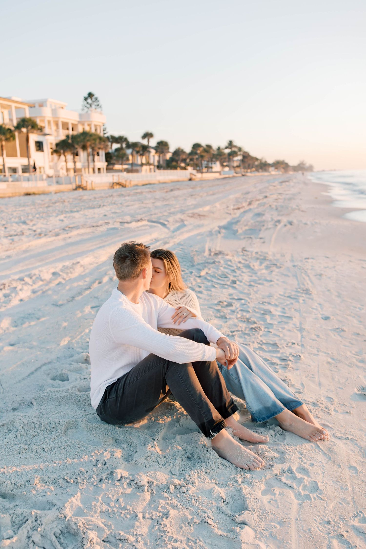 Couple sitting close together on white sandy beach during golden hour.
