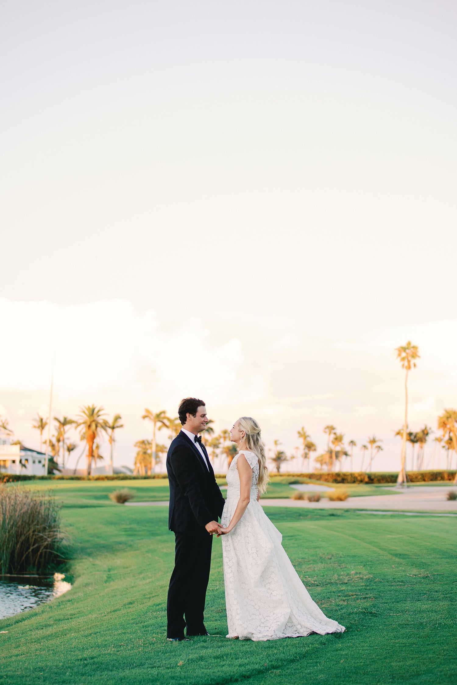 Bride and groom hold hands on lush green golf course at sunset with palm trees in background.