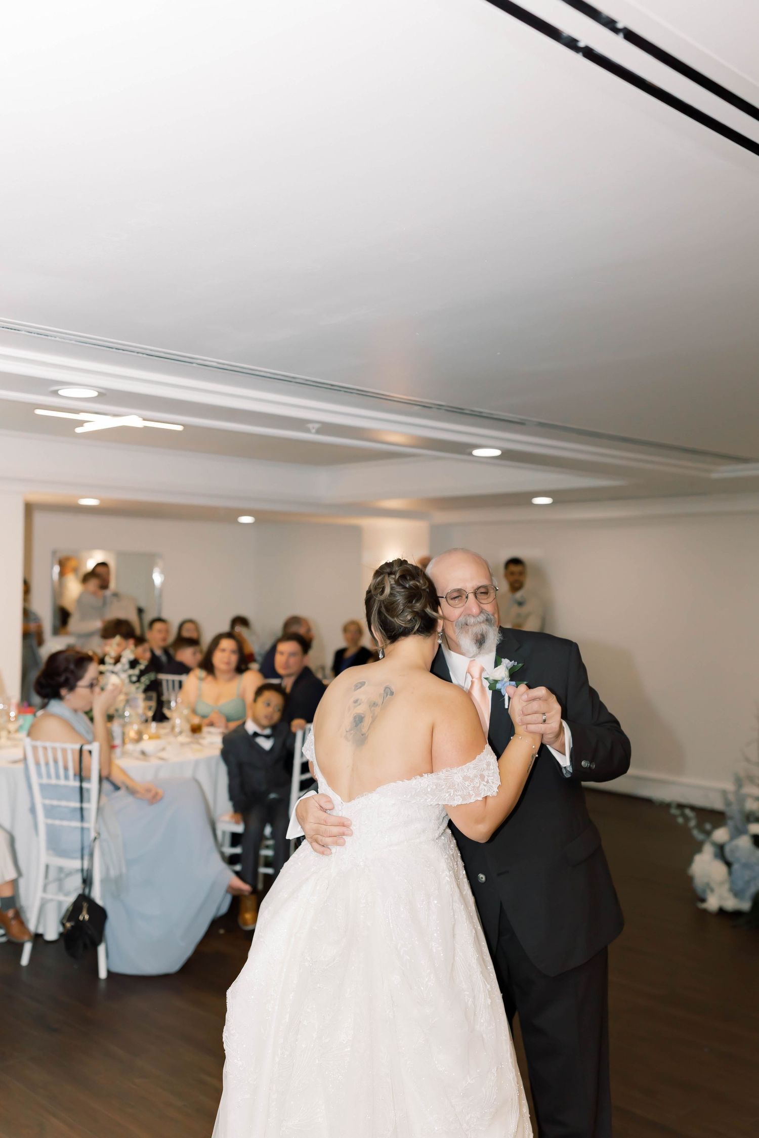 Bride with back tattoo shares emotional father-daughter dance at elegant wedding reception with seated guests.