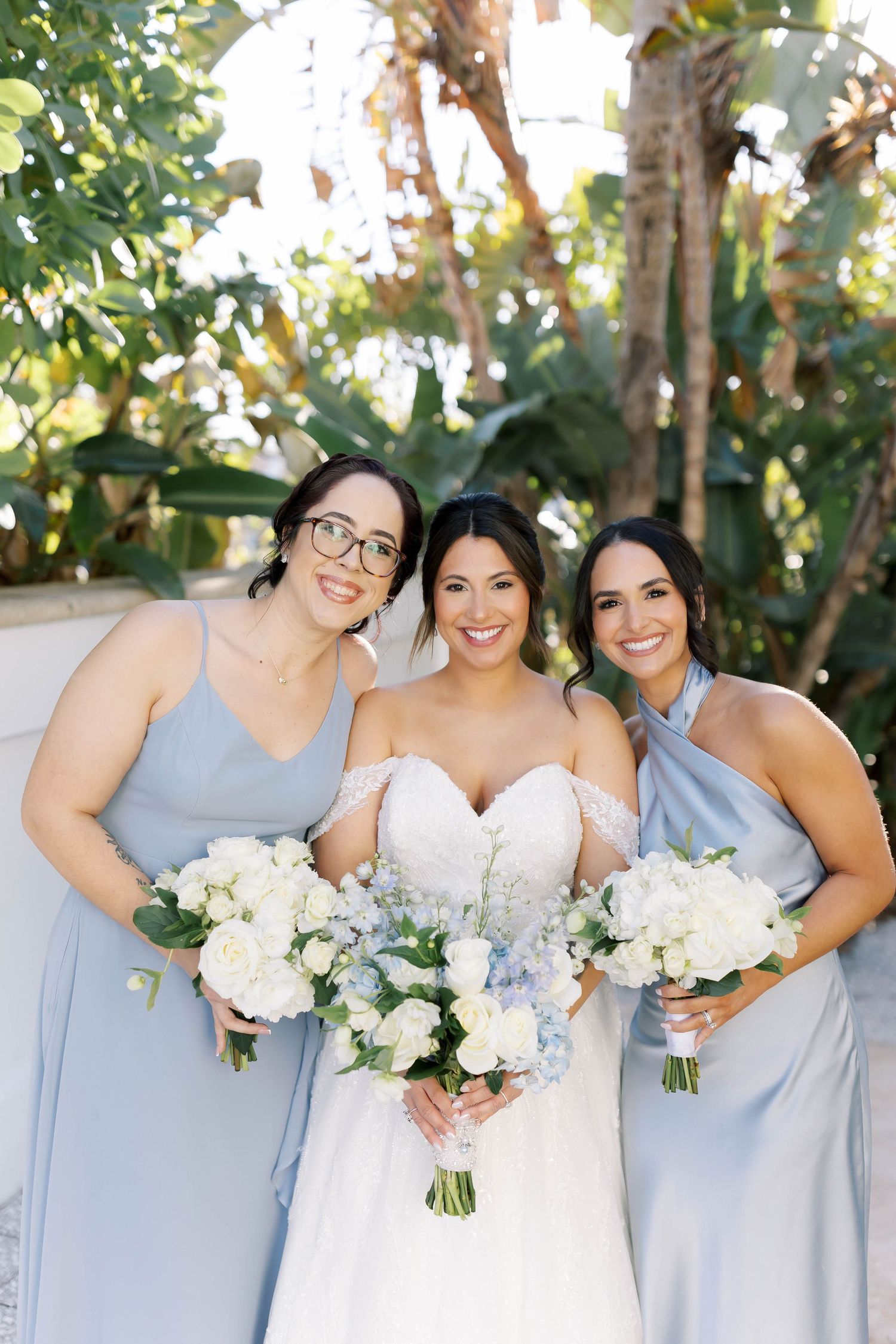 Bride in white lace gown poses with two bridesmaids in light blue dresses holding white and blue bouquets outdoors.