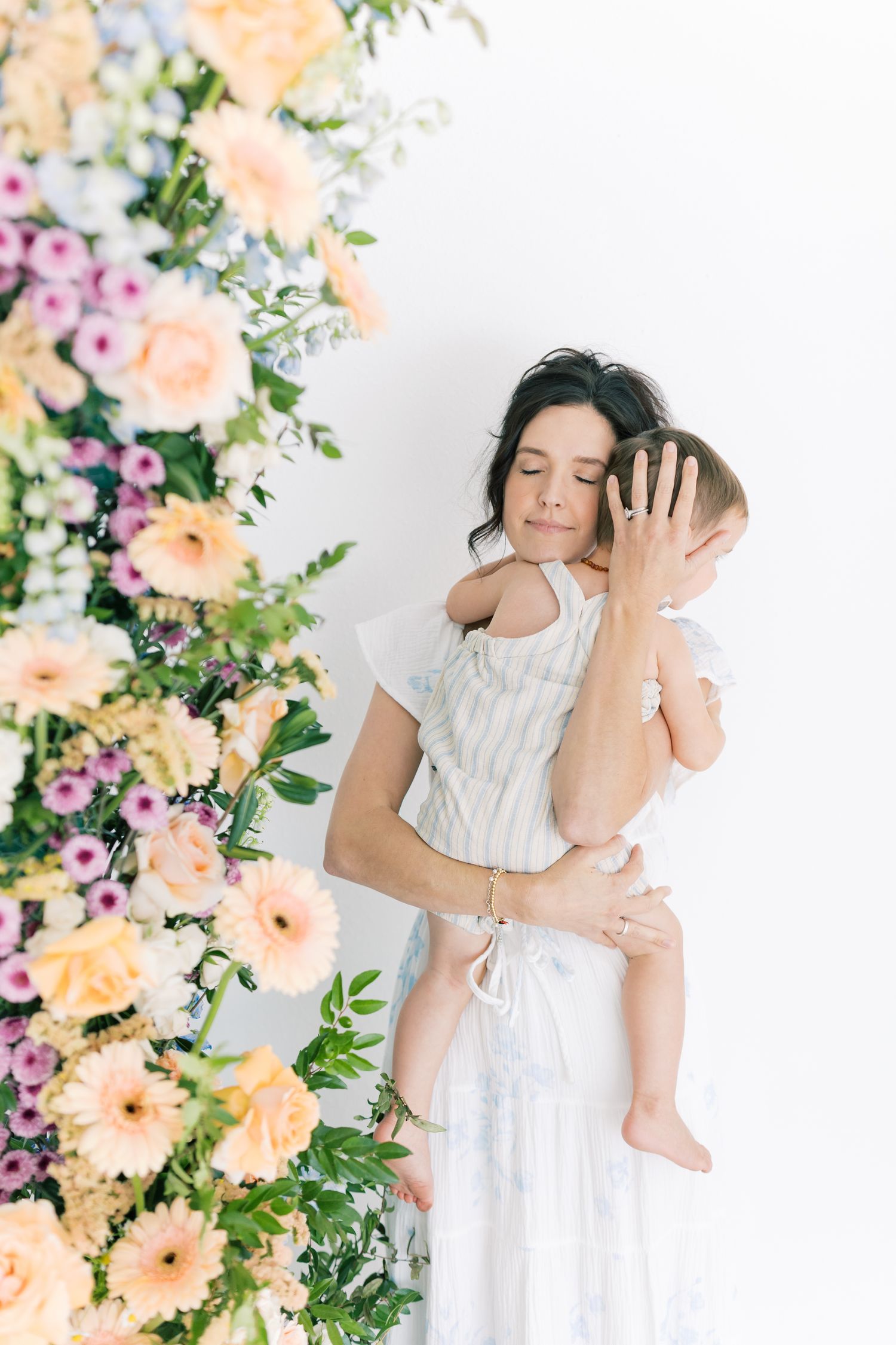 A mother in a white dress tenderly holds her baby near a lush floral arrangement with pastel blooms.
