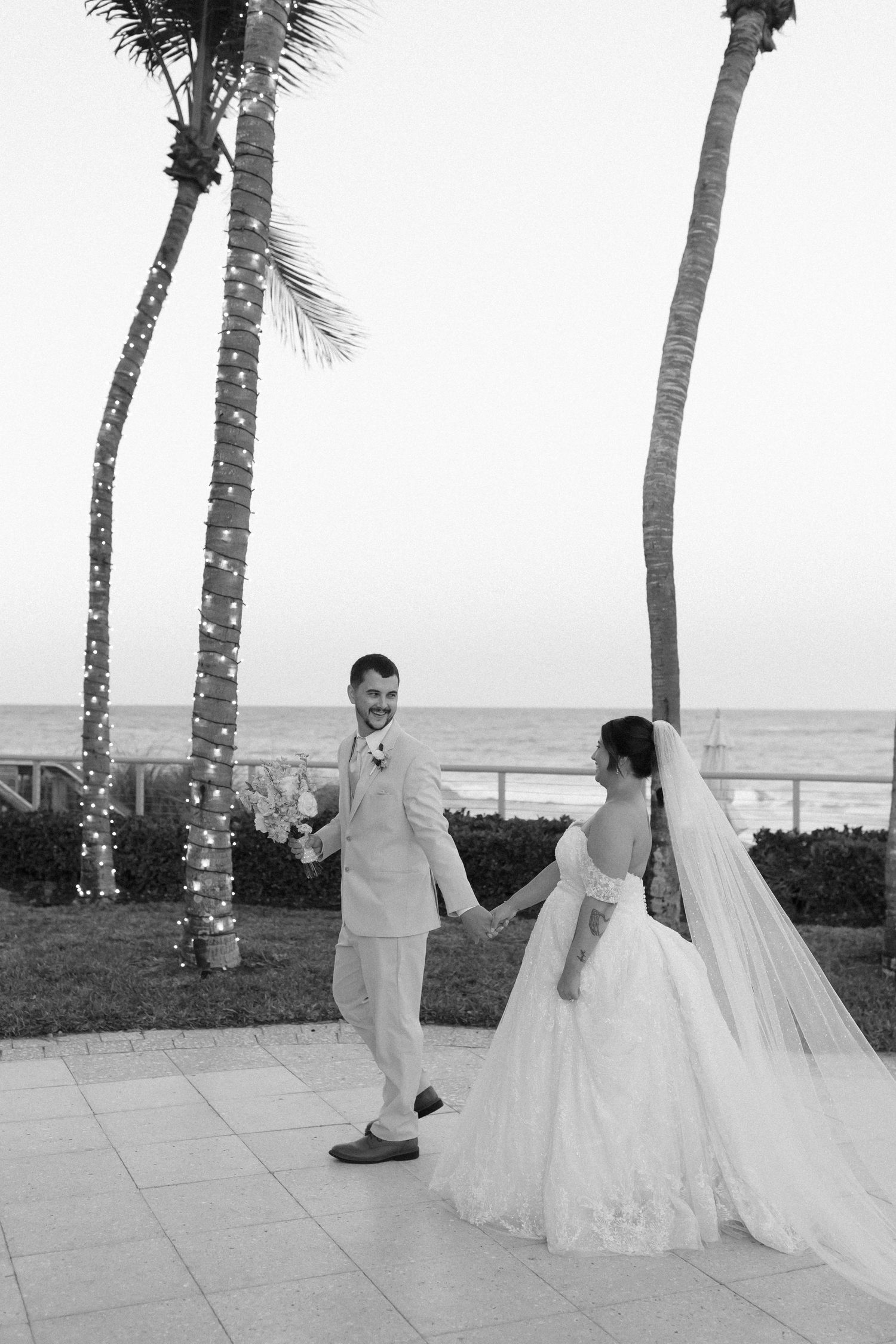 Black and white photo of bride and groom holding hands near palm trees with string lights and ocean backdrop at dusk.