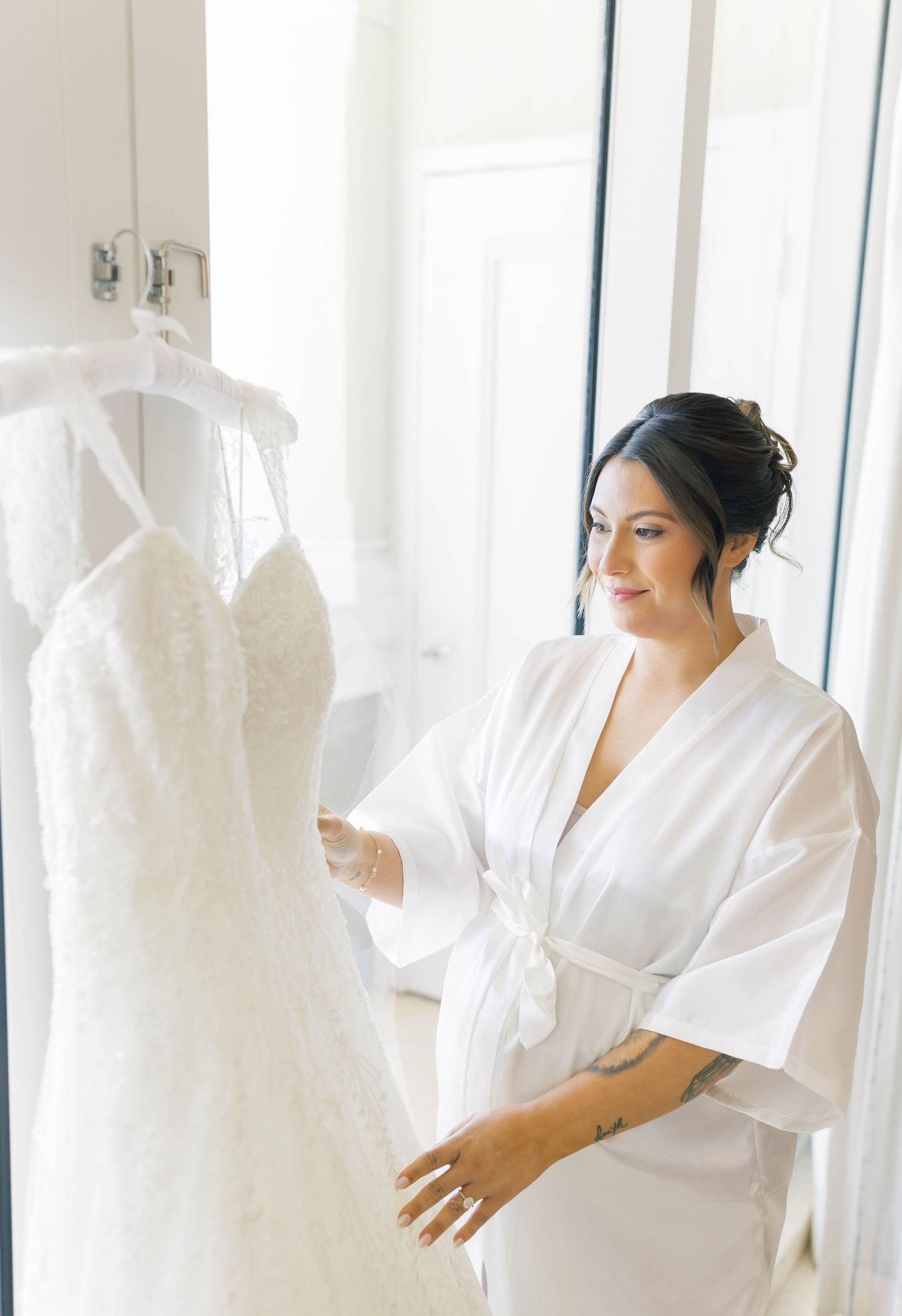 Bride in white robe admiring her lace wedding dress hanging on a door before her wedding ceremony.