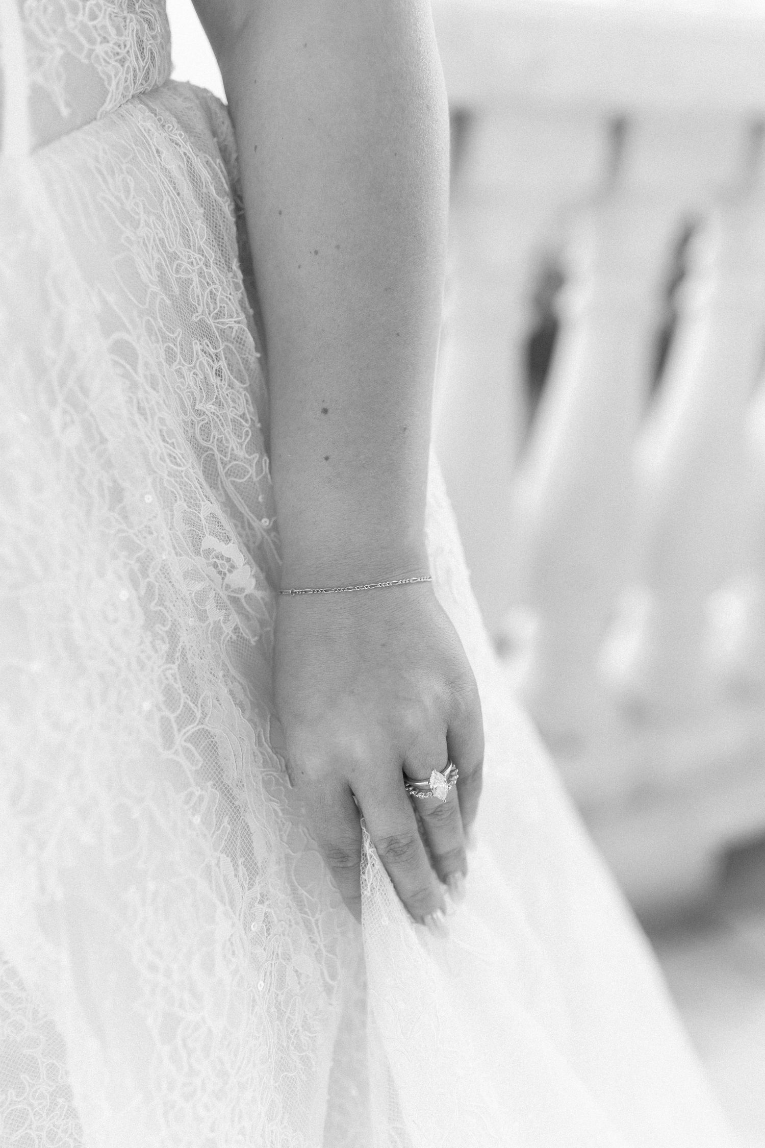Black and white close-up of a bride's hand wearing a diamond engagement ring against a lace wedding dress.
