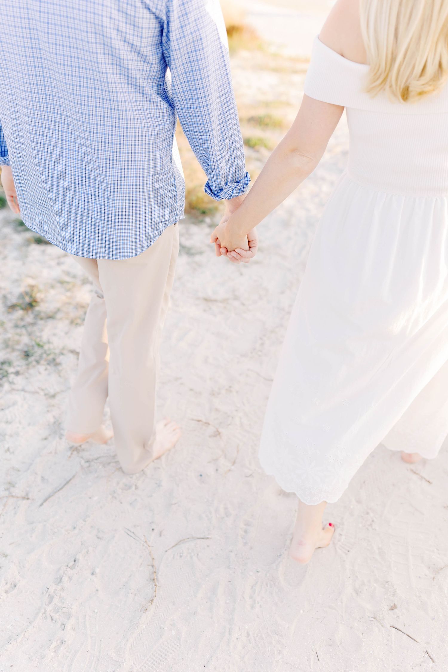 Couple in blue and white outfits walking barefoot on sandy beach at sunset.