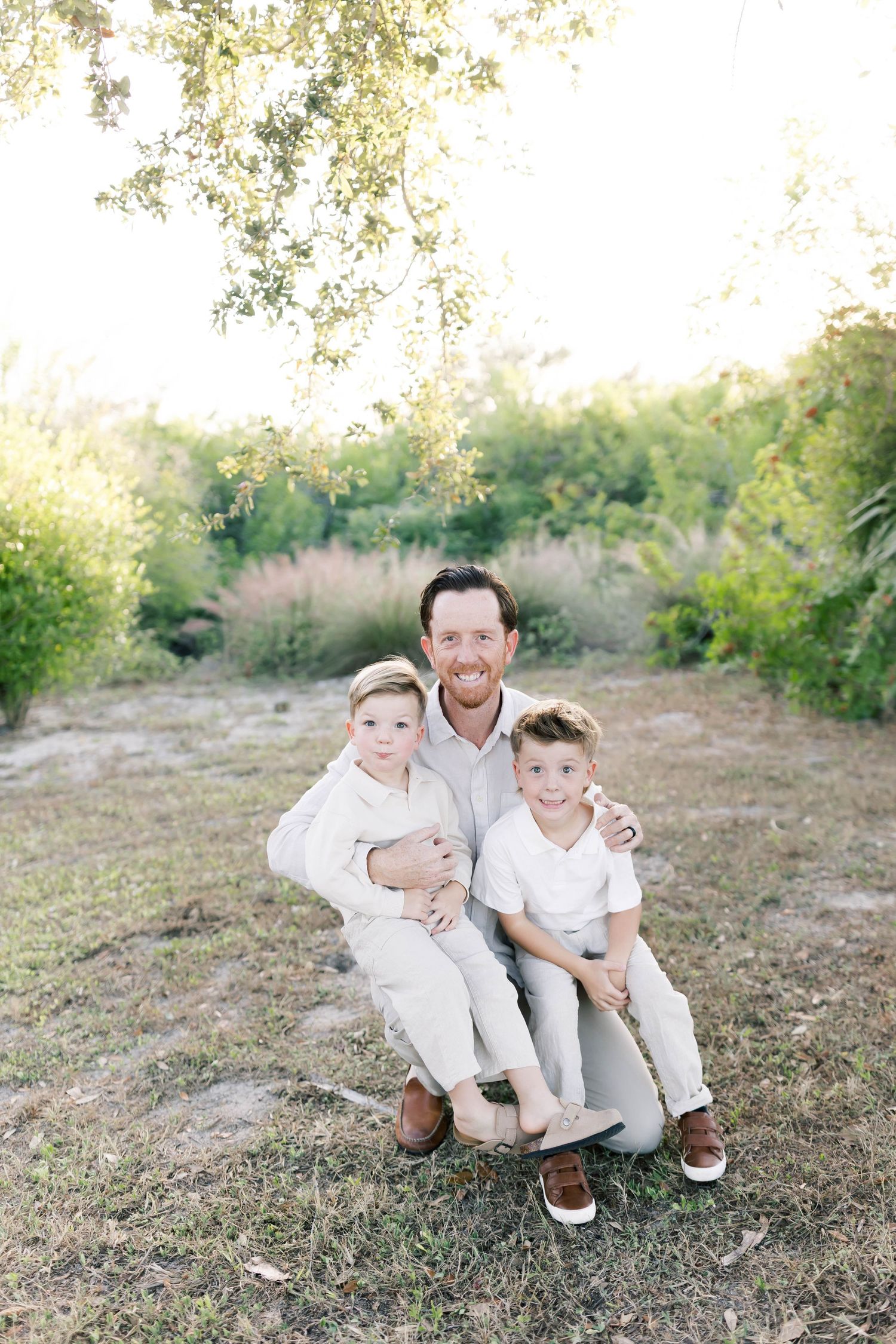 A heartwarming outdoor family portrait in white outfits against a natural backdrop with soft sunlight filtering through trees.