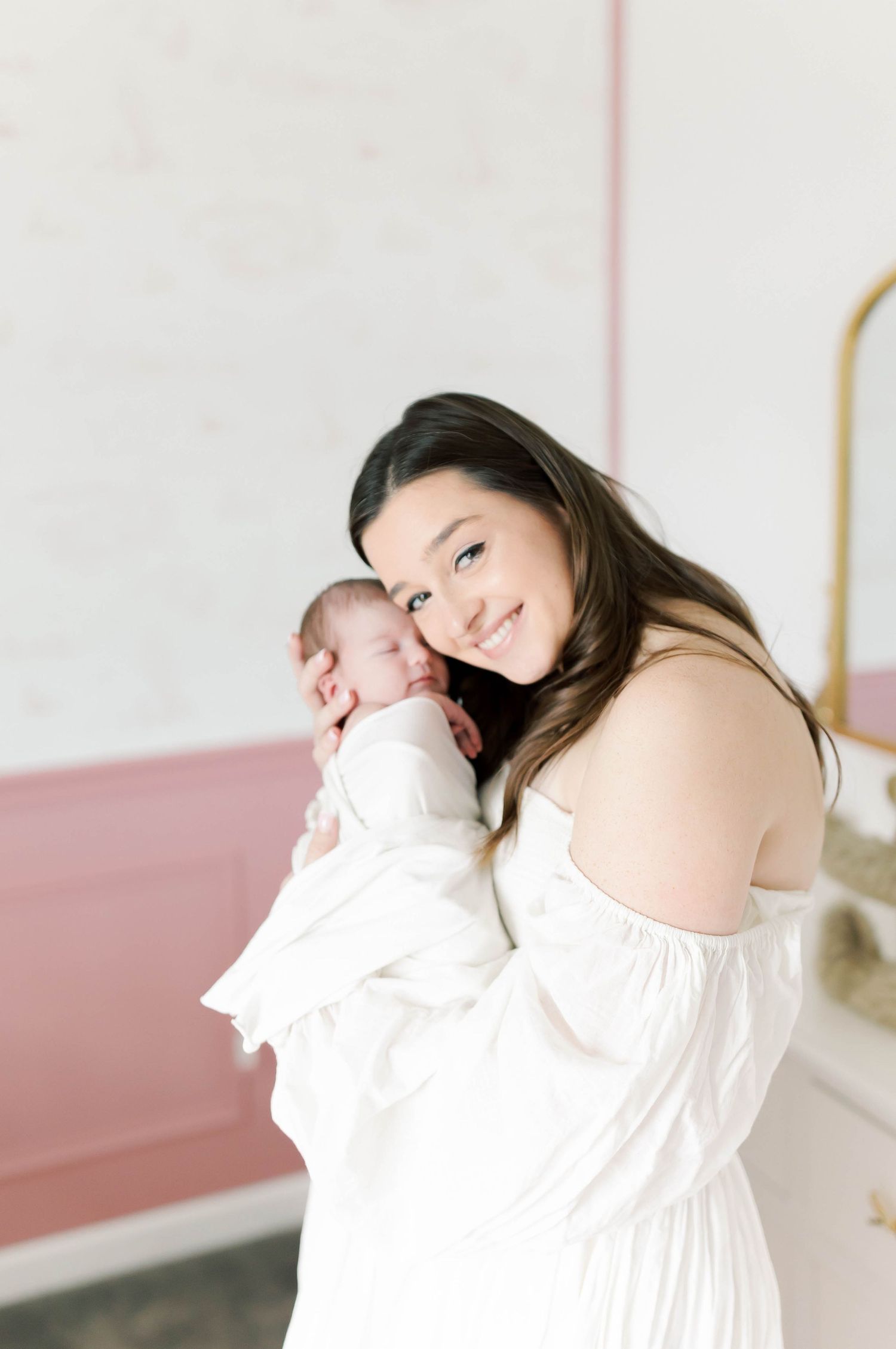 Mother in white dress holding sleeping newborn baby in bright nursery setting.