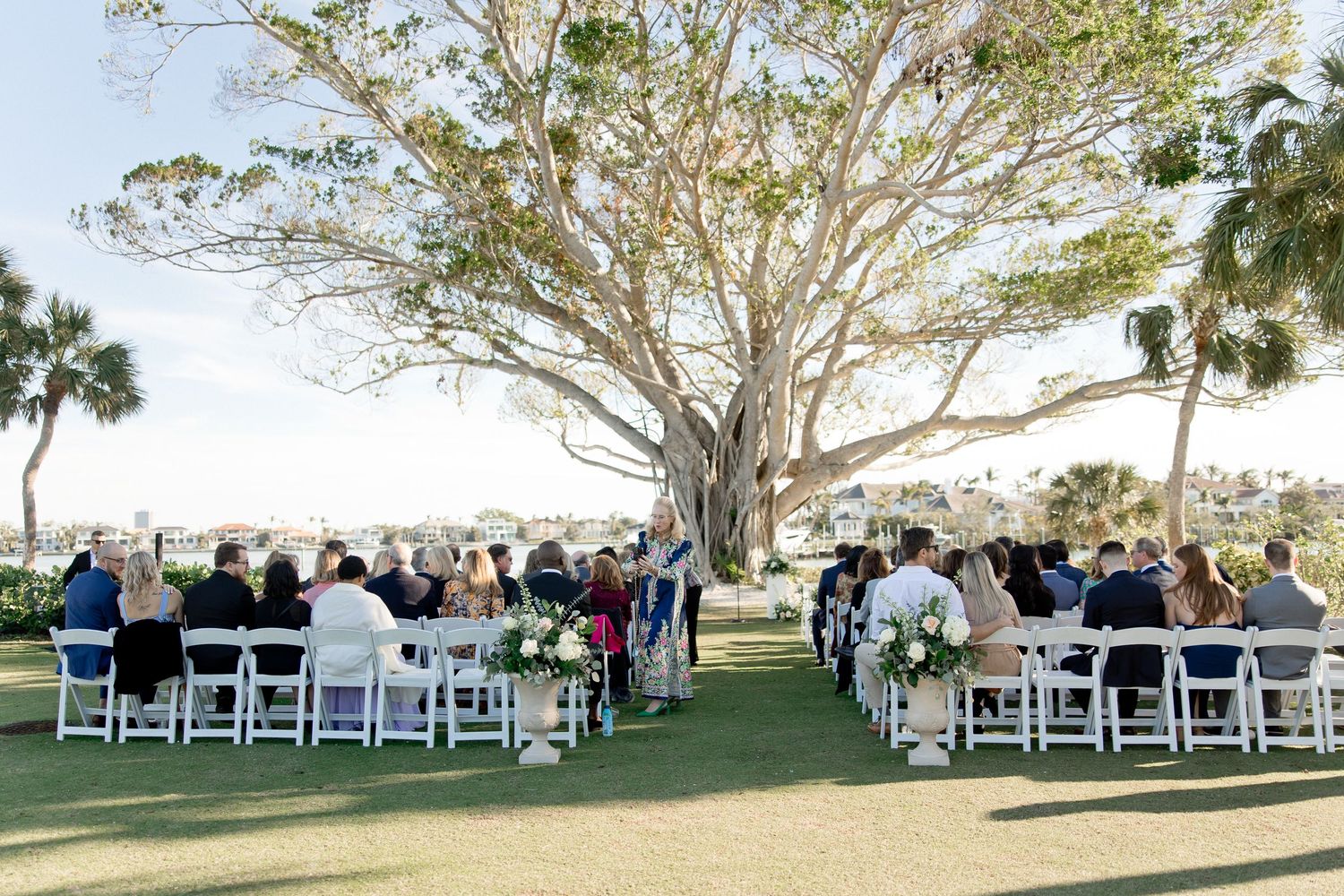 Outdoor wedding ceremony setup with white chairs under trees and natural lighting.