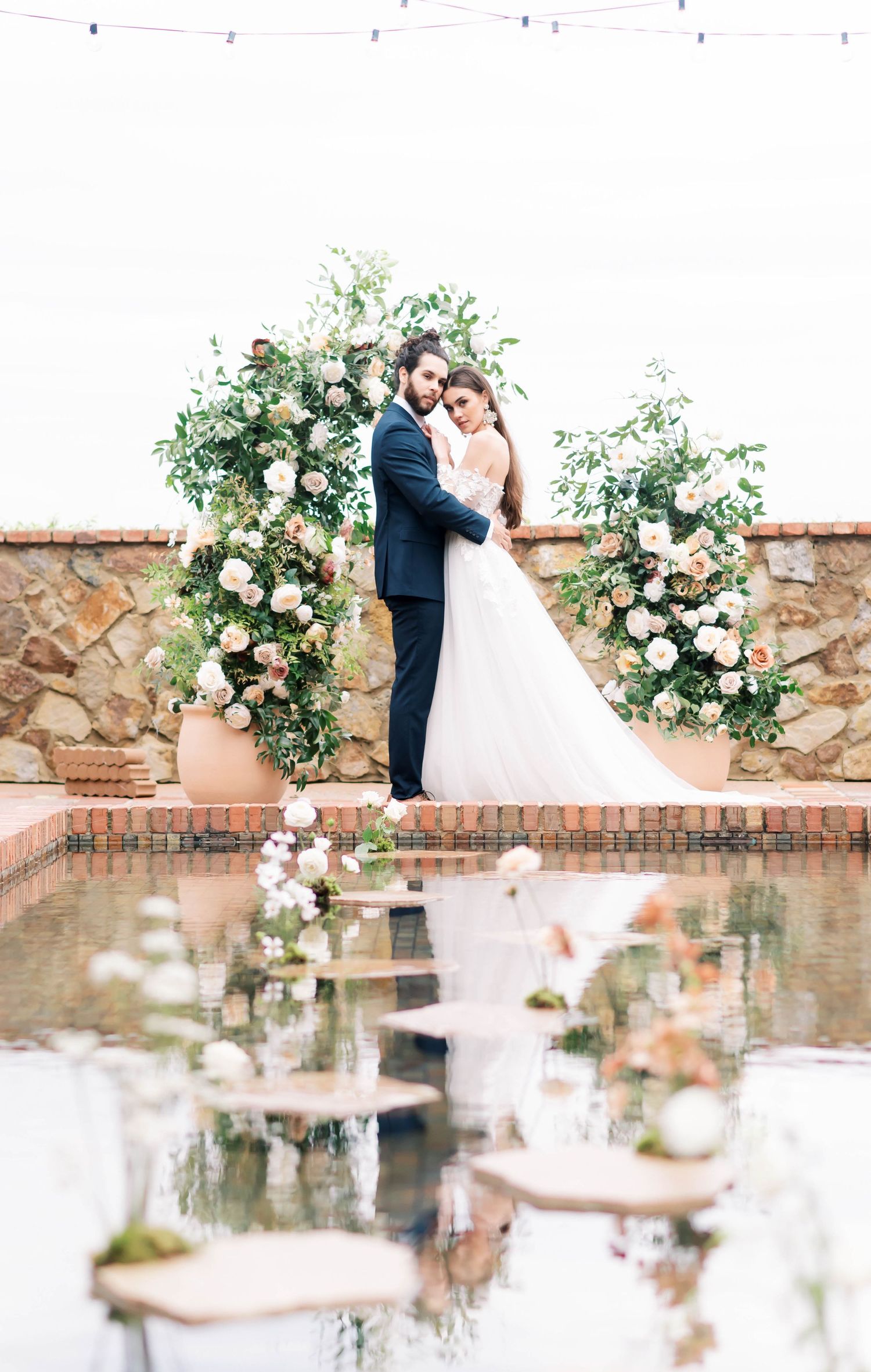 Bride and groom embrace before floral arch with blush roses by reflective pool at elegant outdoor wedding venue.