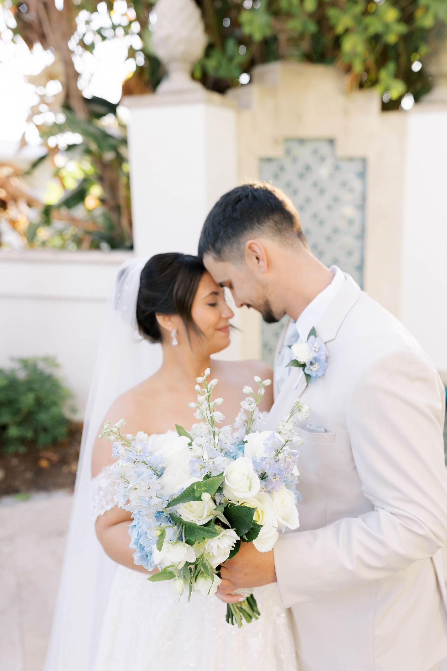 Bride and groom share a romantic forehead touch, bride holding a blue and white floral bouquet outdoors.