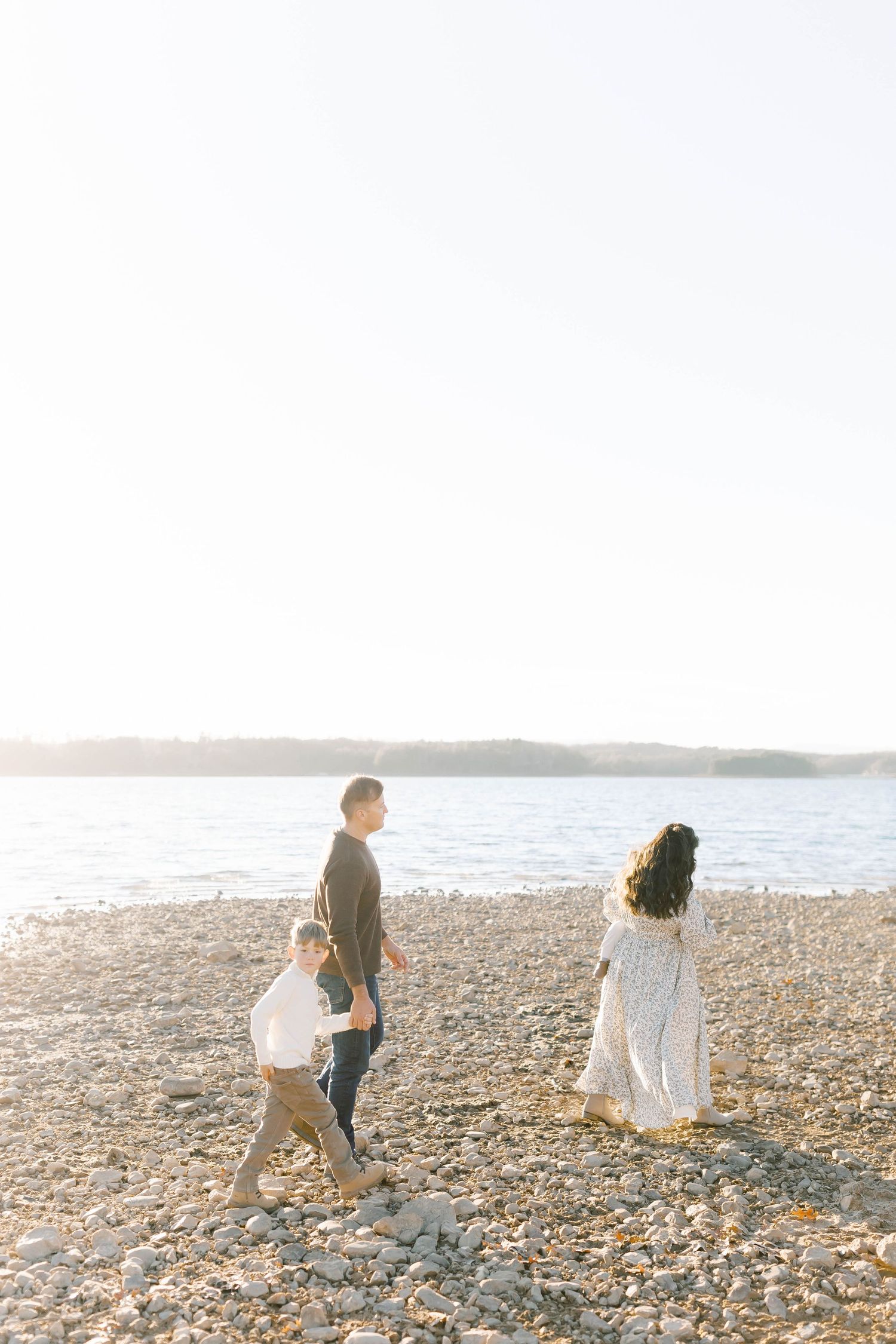 A family walks together on a rocky beach at sunset with water and trees in the background.
