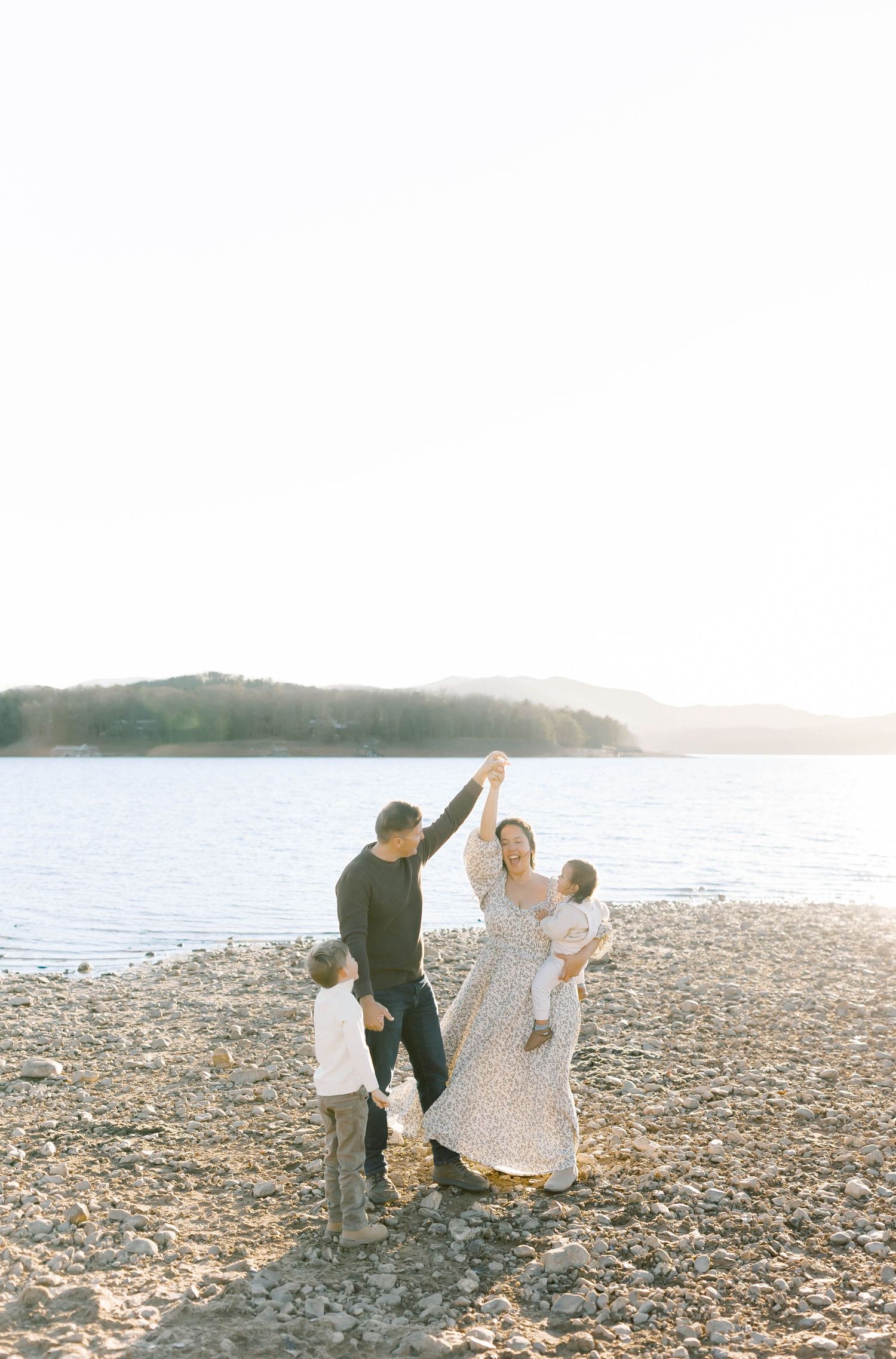 A joyful family dances together on a pebble beach with a serene lake view.