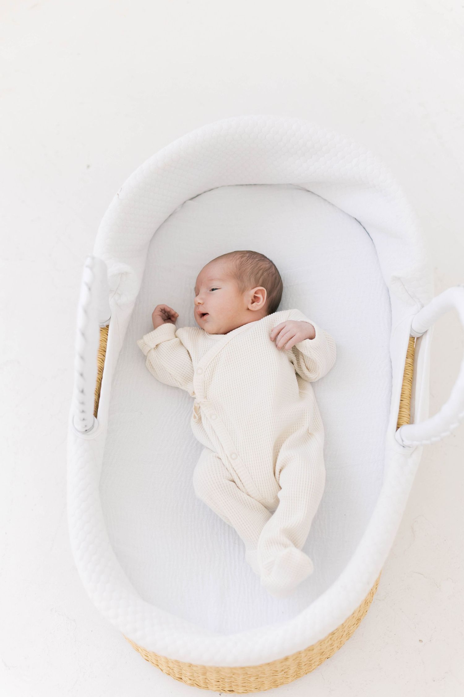 A sleeping newborn baby wearing cream colored clothing rests peacefully in a white bassinet.