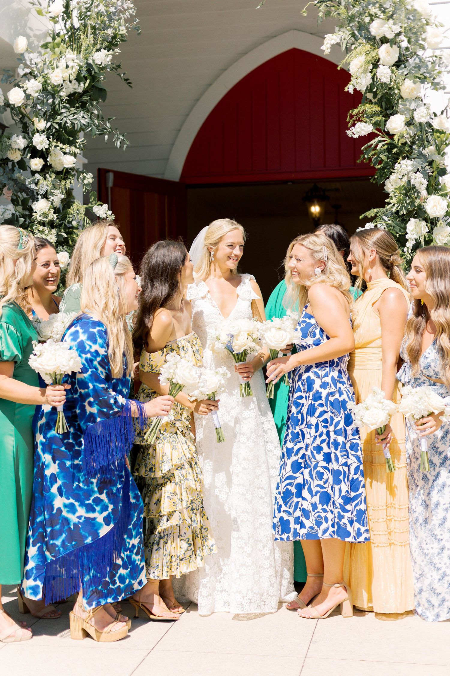 Bridal party in colorful floral dresses gathered around bride under floral arch entrance.