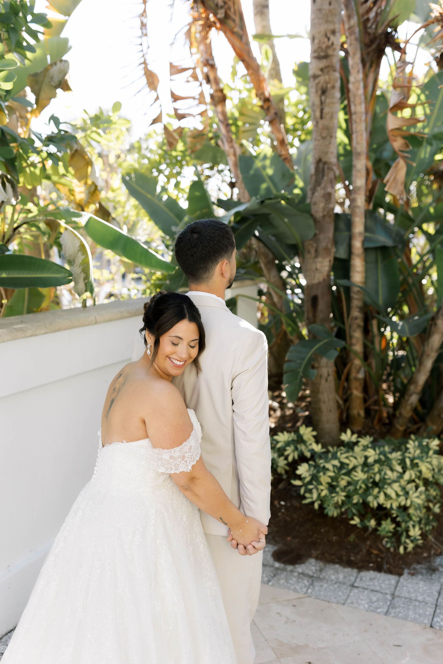 Bride in off-shoulder gown holds groom's hand, smiling among lush tropical greenery at outdoor wedding venue.