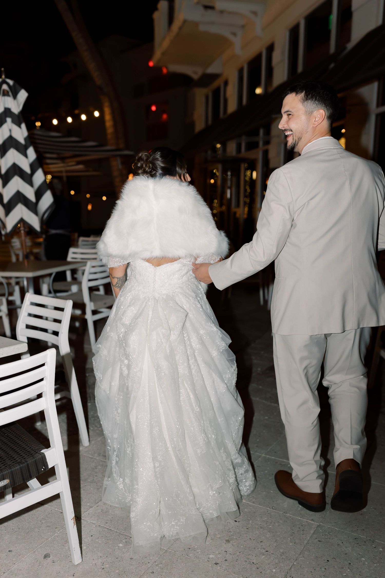 Groom in beige suit escorts bride in white fur wrap and lace gown through an outdoor evening reception venue.