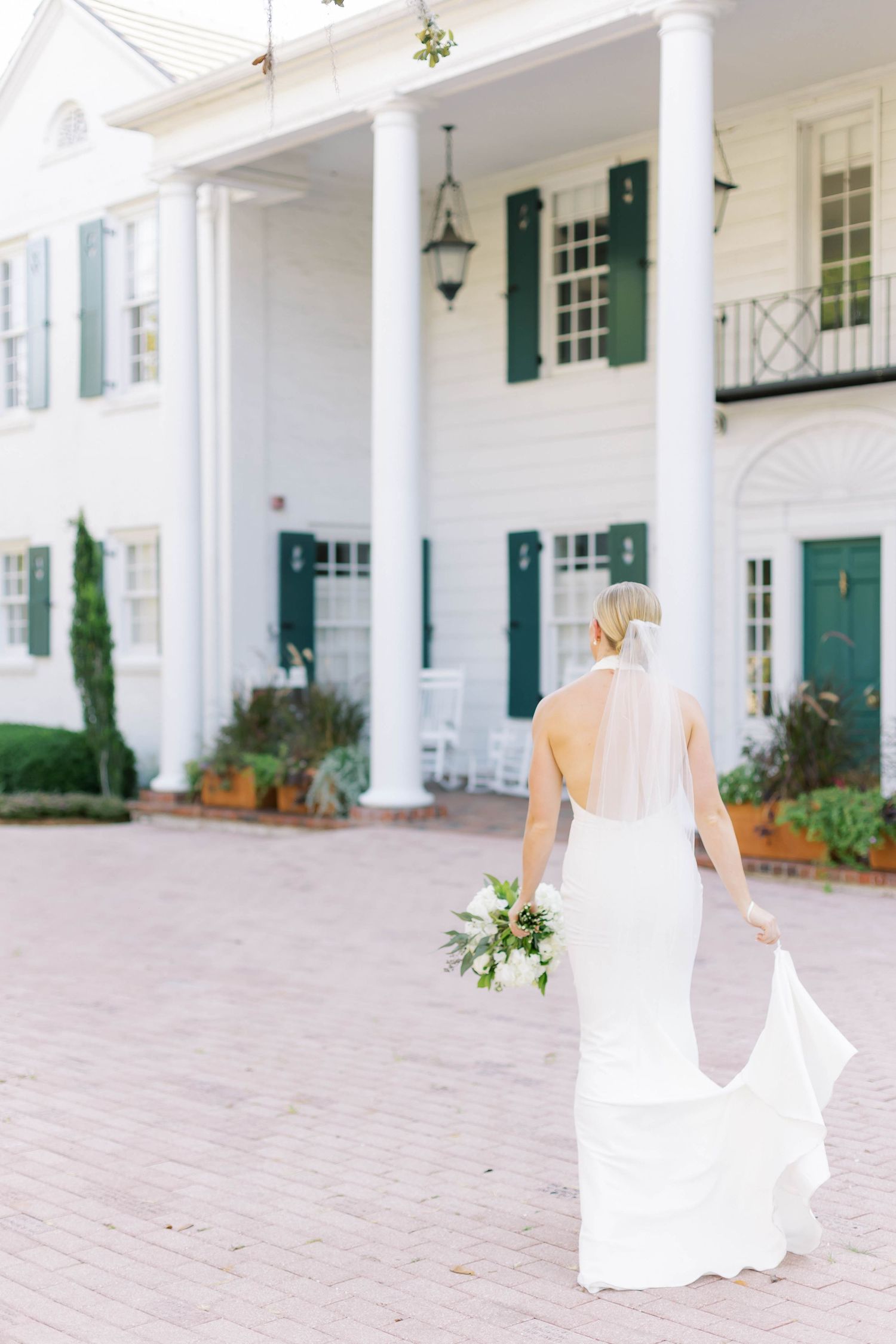 Bride in white gown with veil holds bouquet walking toward elegant white colonial venue with green shutters.