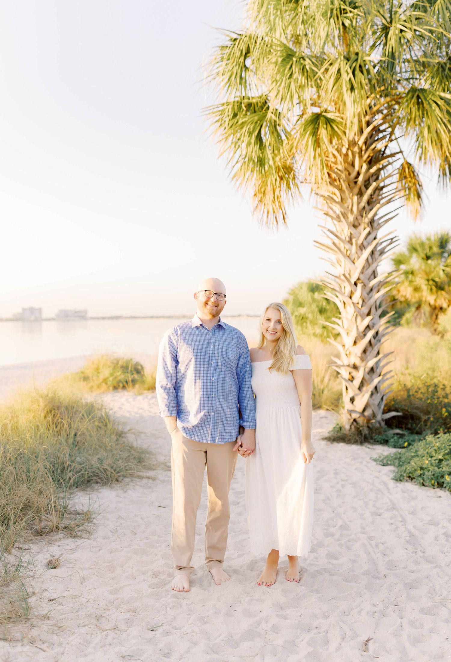 Couple walking barefoot on sandy path with palm trees in background.