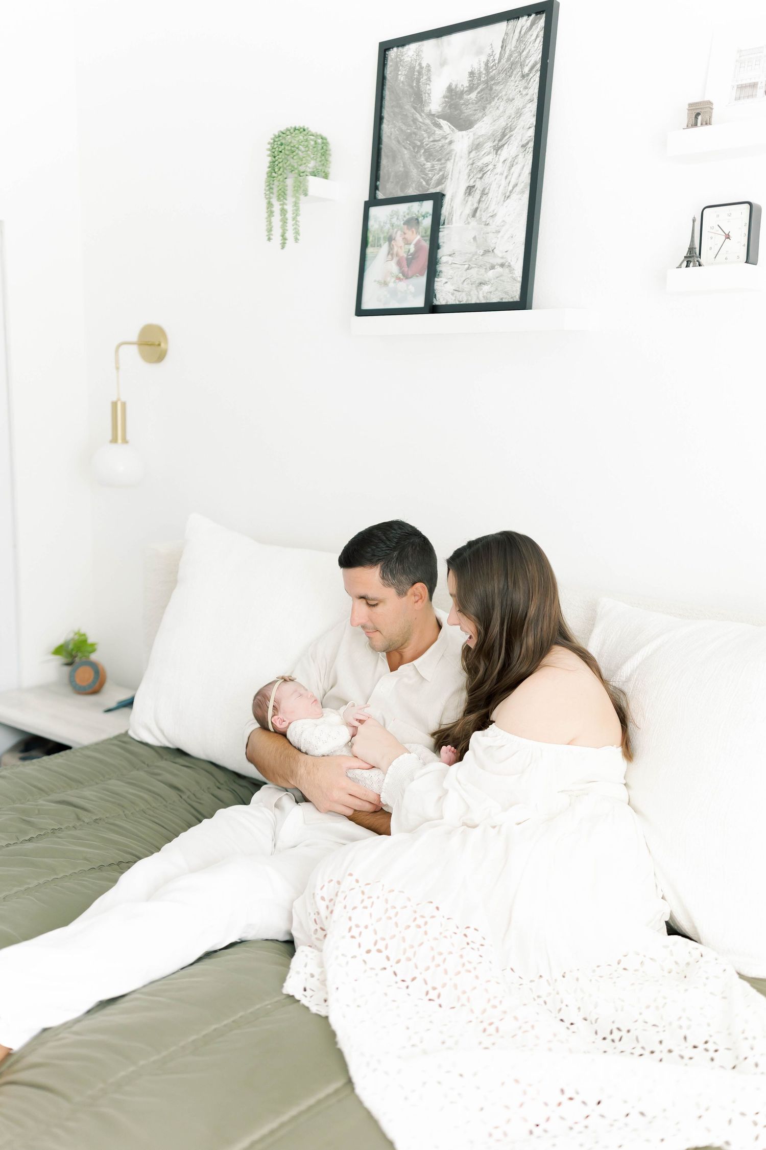 Parents sitting on bed with newborn baby in bright modern bedroom with hanging plants.