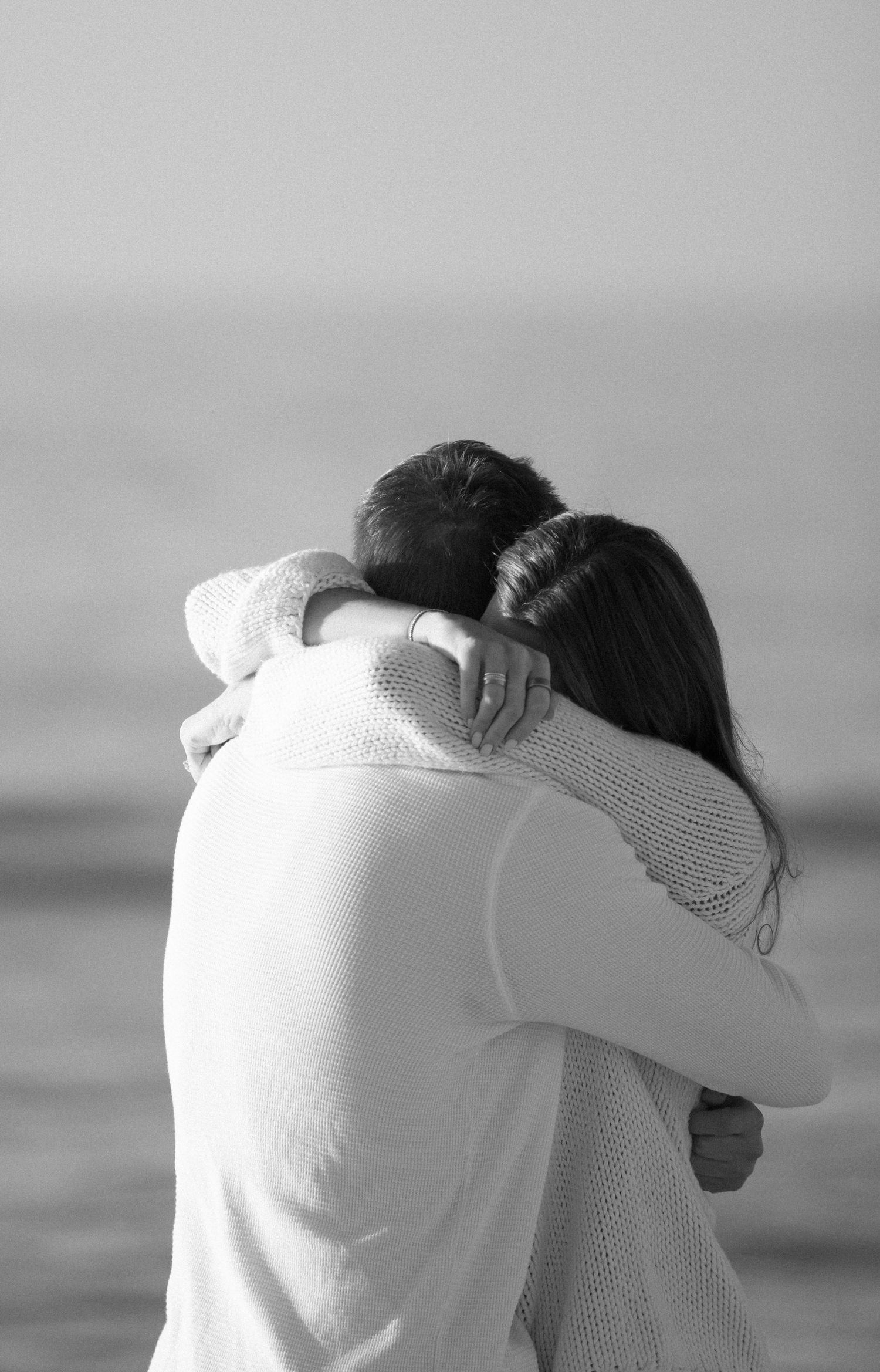 A tender embrace between two people wearing white sweaters against an ocean backdrop.