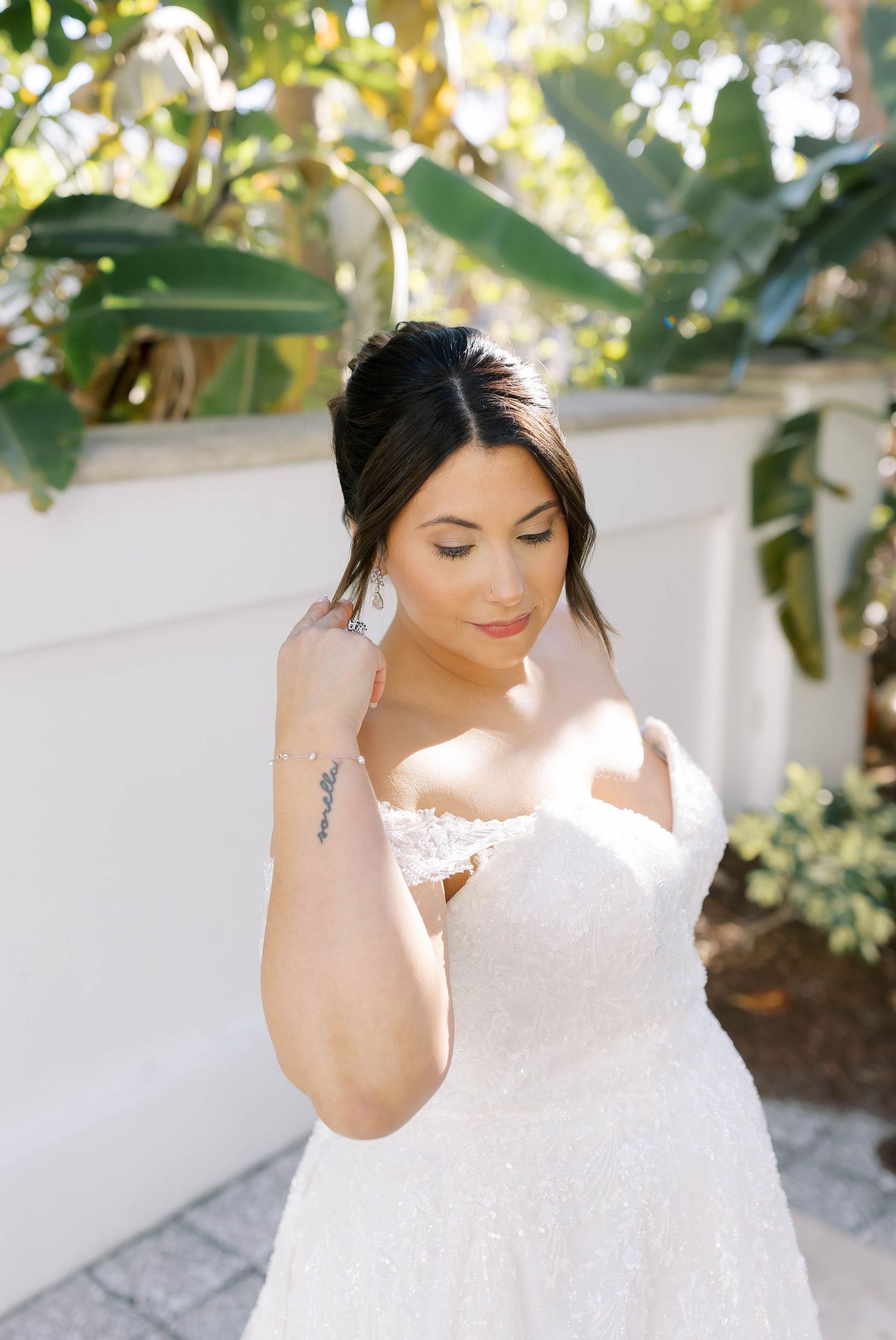 Brunette bride in strapless lace wedding gown adjusts earring surrounded by tropical greenery.