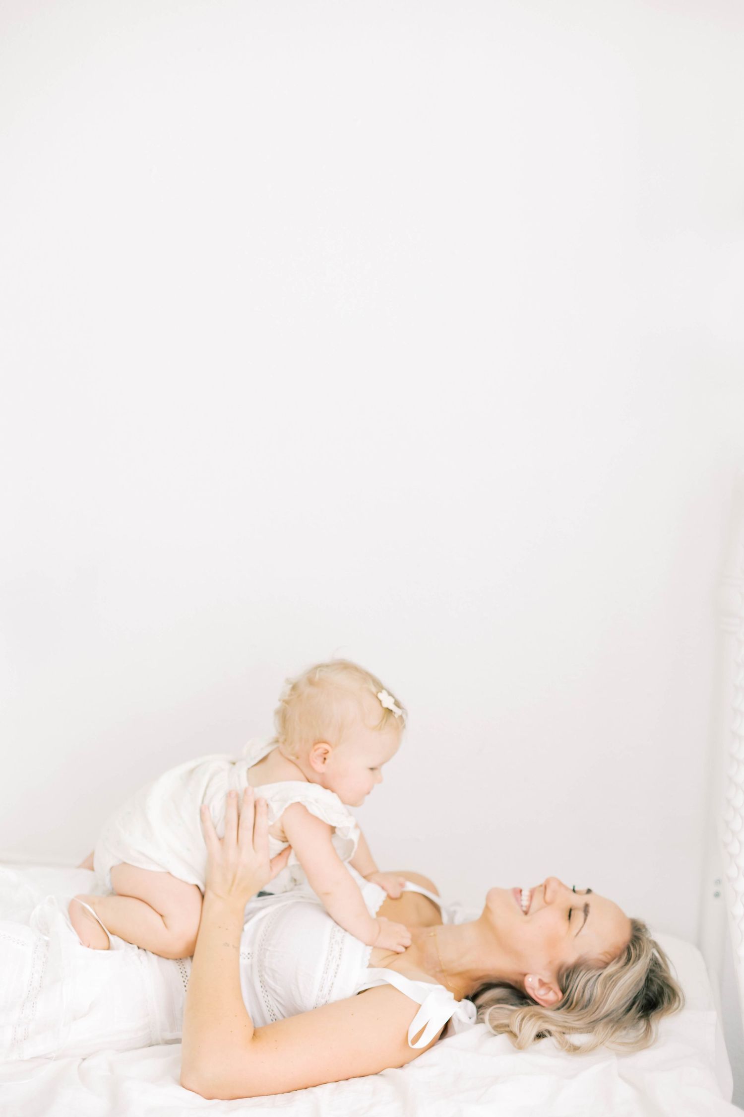 Mother lying on white bed holding her baby girl up, both dressed in white, sharing a tender moment.
