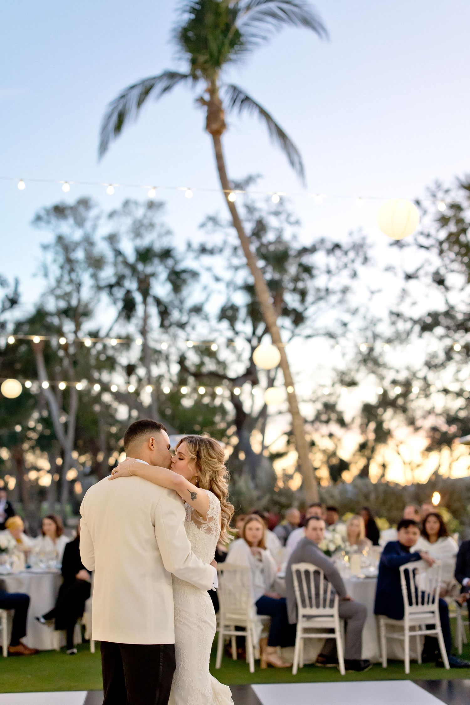 Outdoor wedding reception under palm trees with string lights at sunset.
