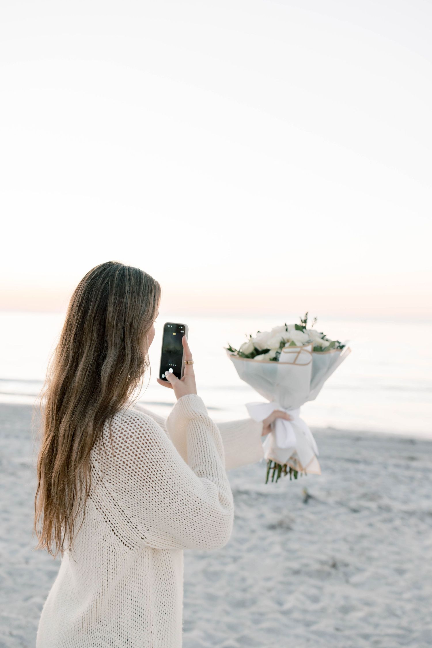 Person in white sweater taking photos of flowers on beach at sunset.
