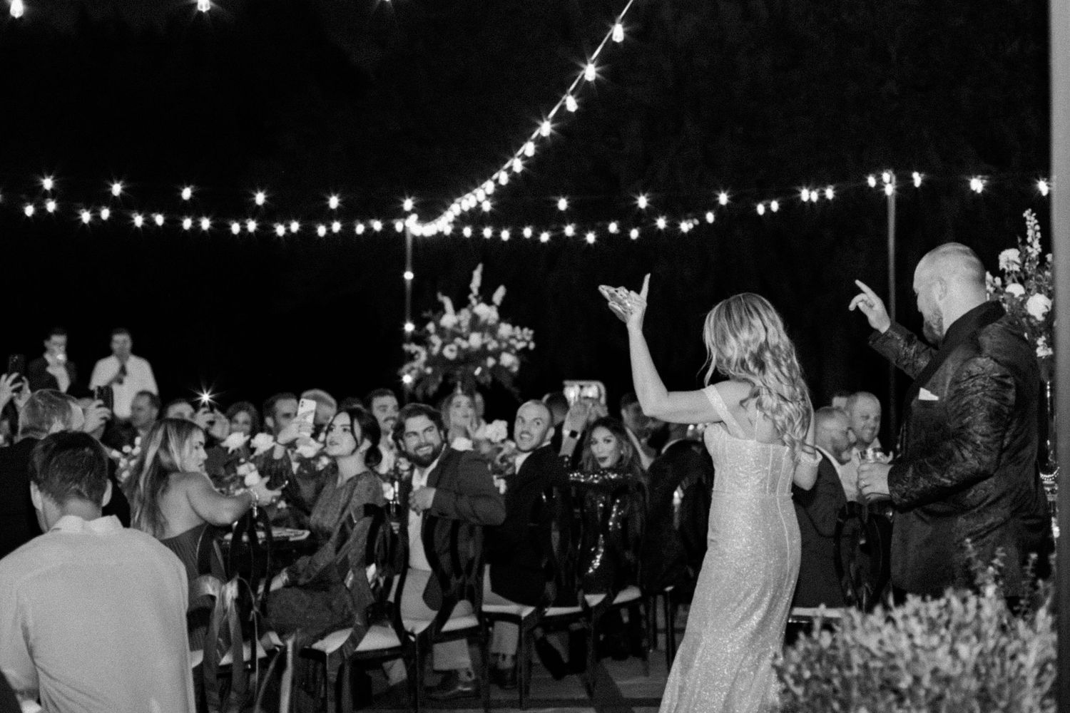 Black and white photo of wedding reception with string lights and dancing guests.