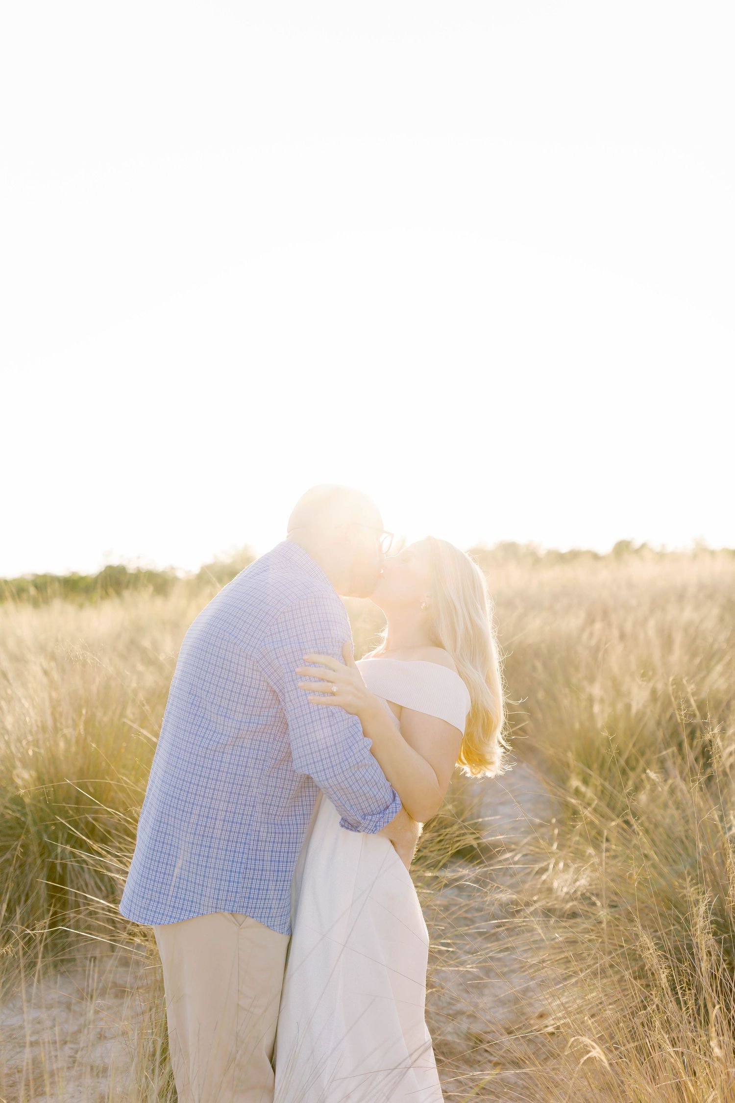 Tender moment between couple in white and blue attire against natural beach grass backdrop.
