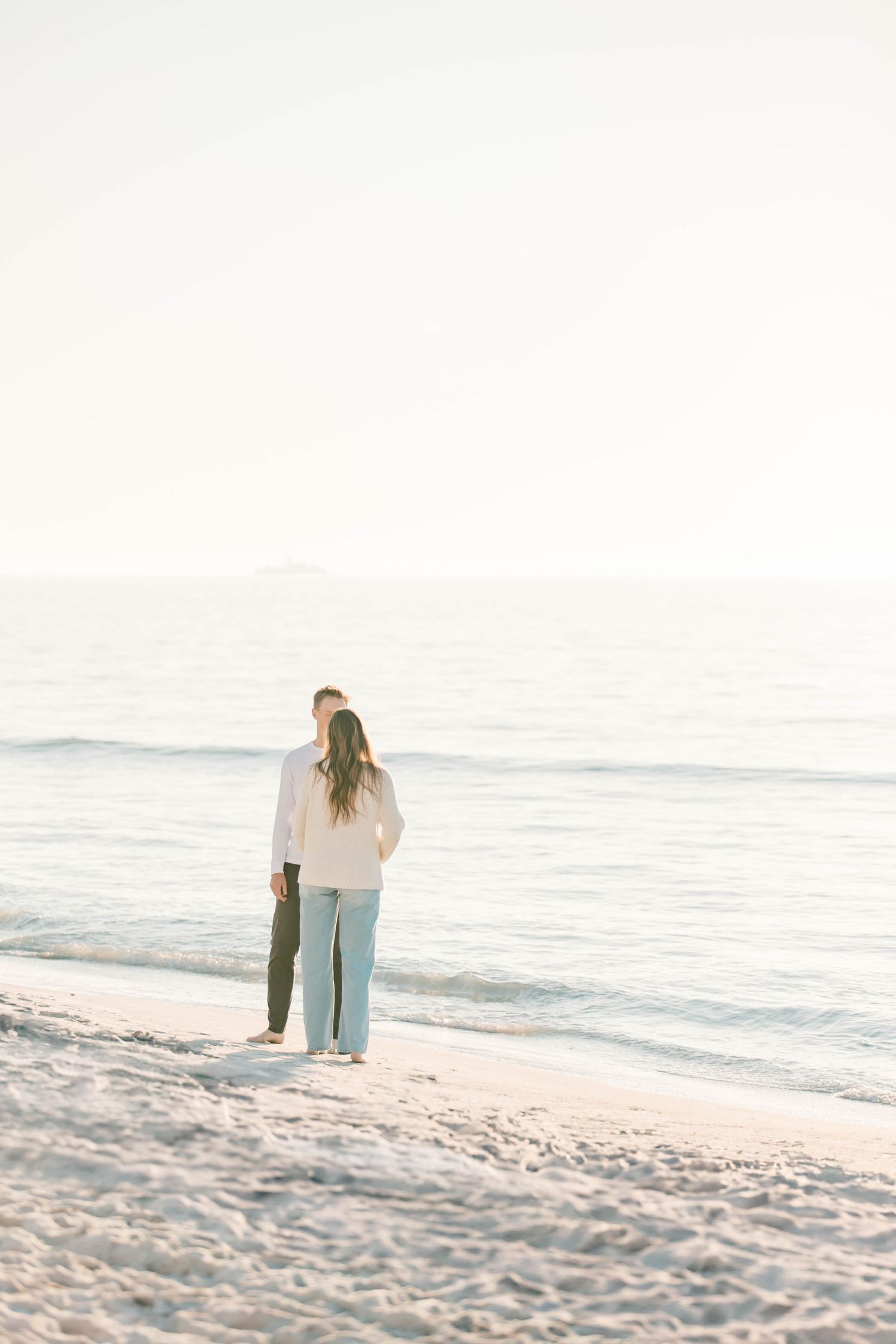 A couple stands together looking out at the ocean on a pristine white sand beach.