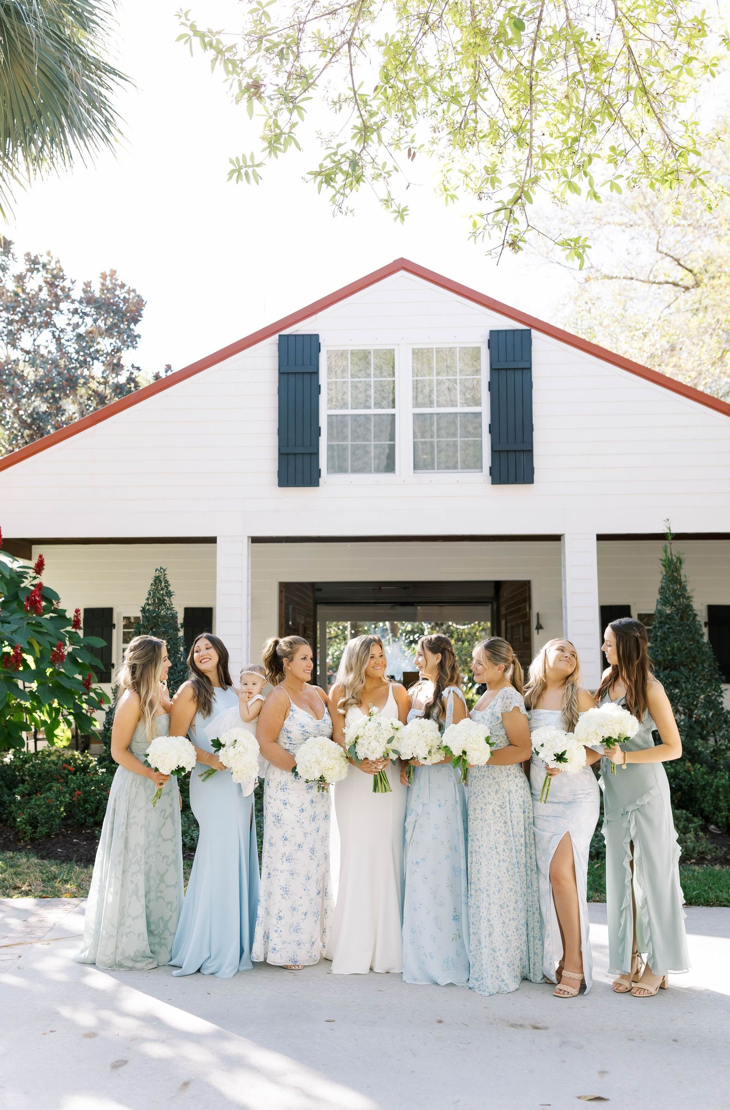 Bridesmaids in light blue dresses pose in front of a white building with navy shutters.