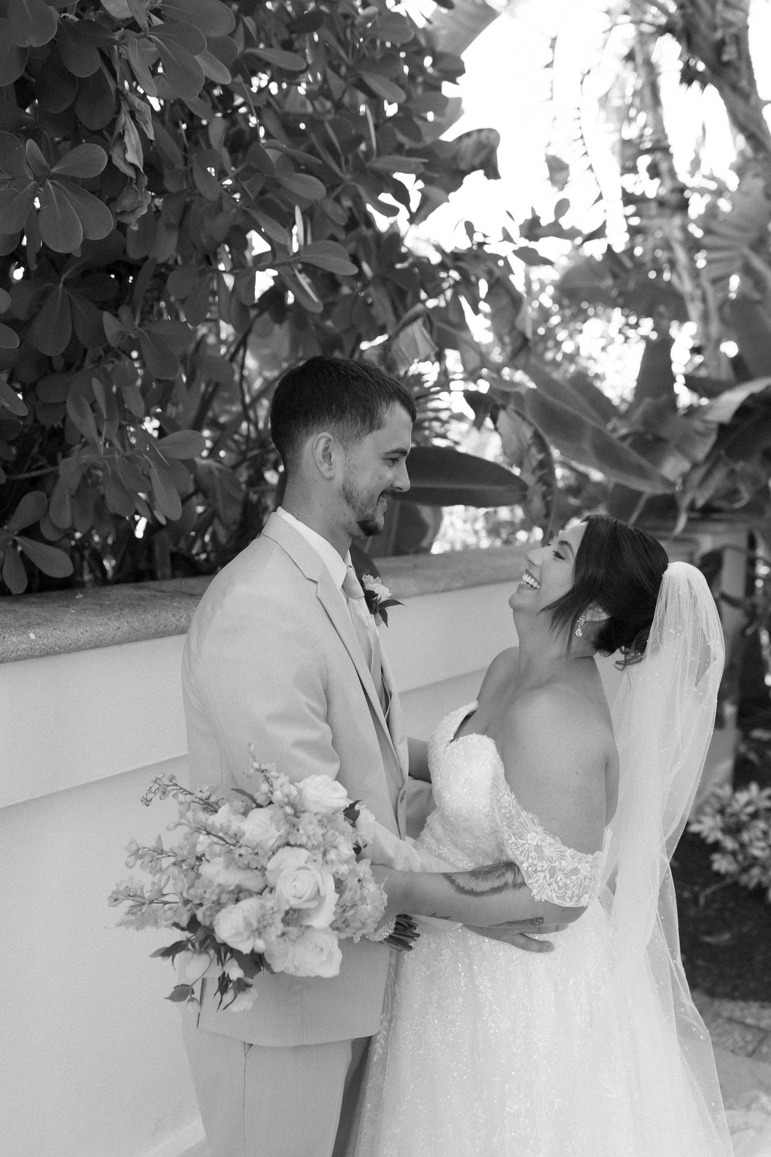 Black and white photo of a smiling bride and groom holding a floral bouquet outdoors on their wedding day.