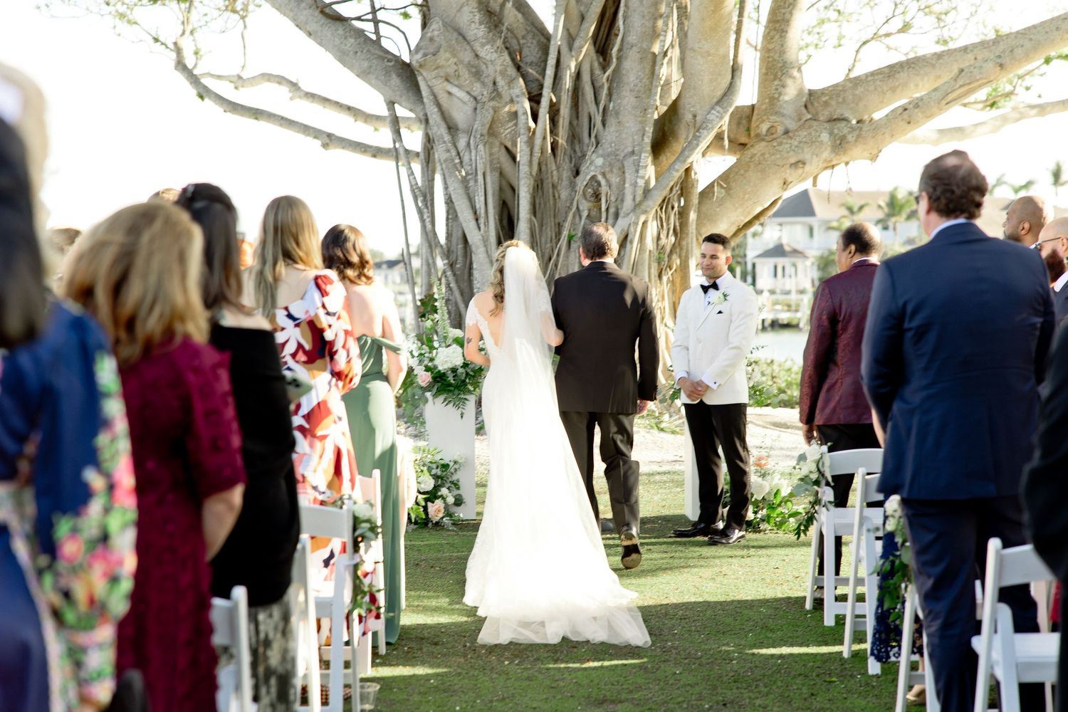 Bride walks down outdoor aisle toward groom under large tree at elegant garden wedding ceremony with seated guests.