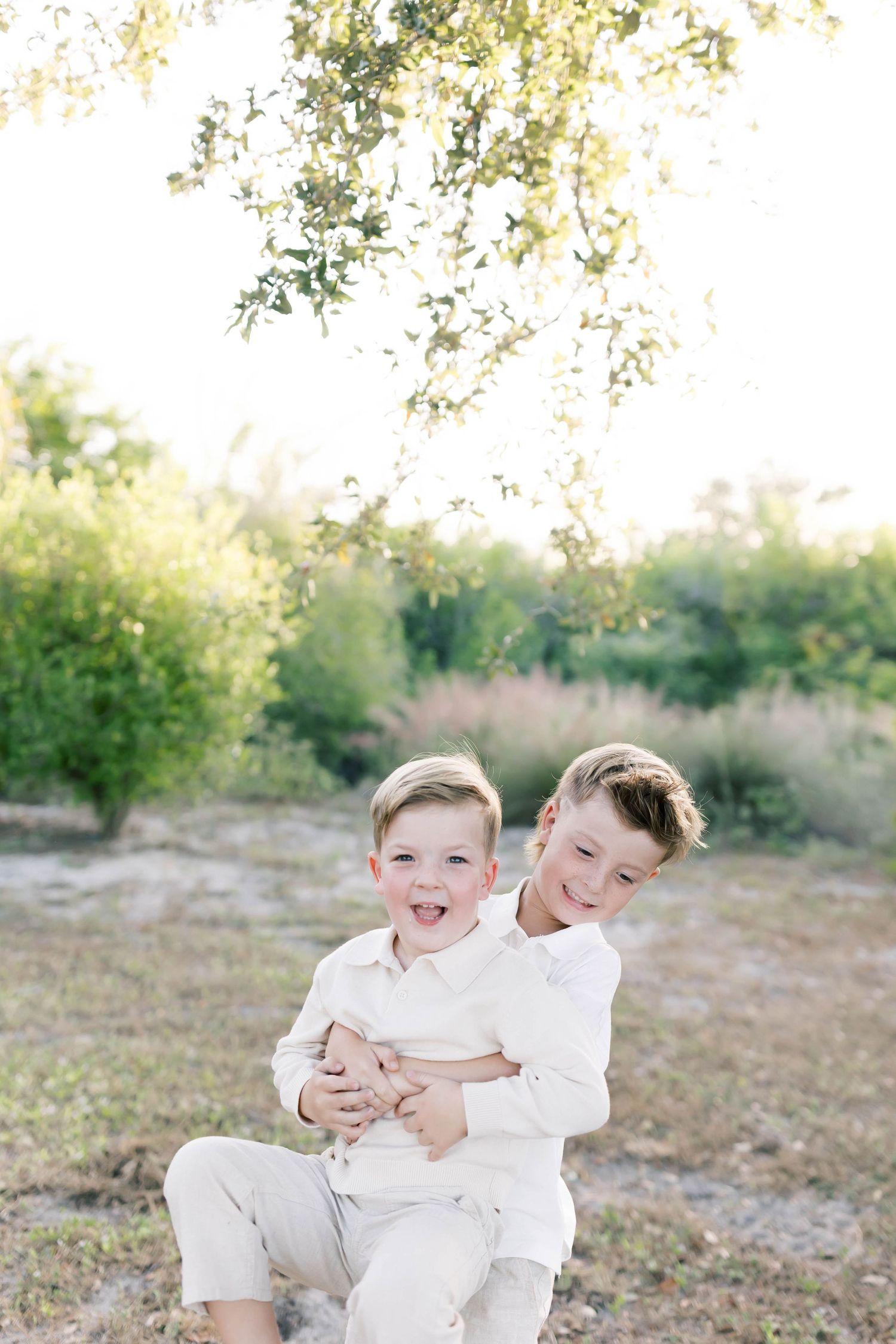Two young siblings in white clothing share a playful embrace while sitting on grass outdoors.