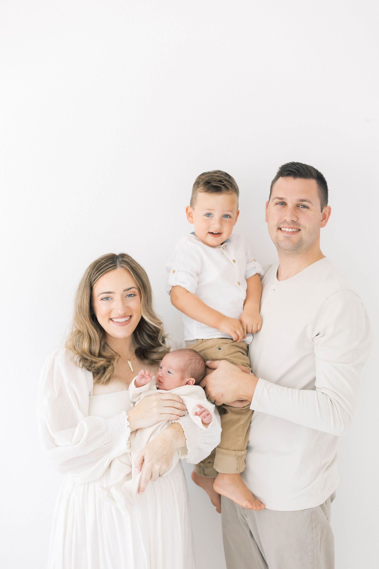Family portrait in white and neutral tones showing tender moments with their newborn baby and toddler.