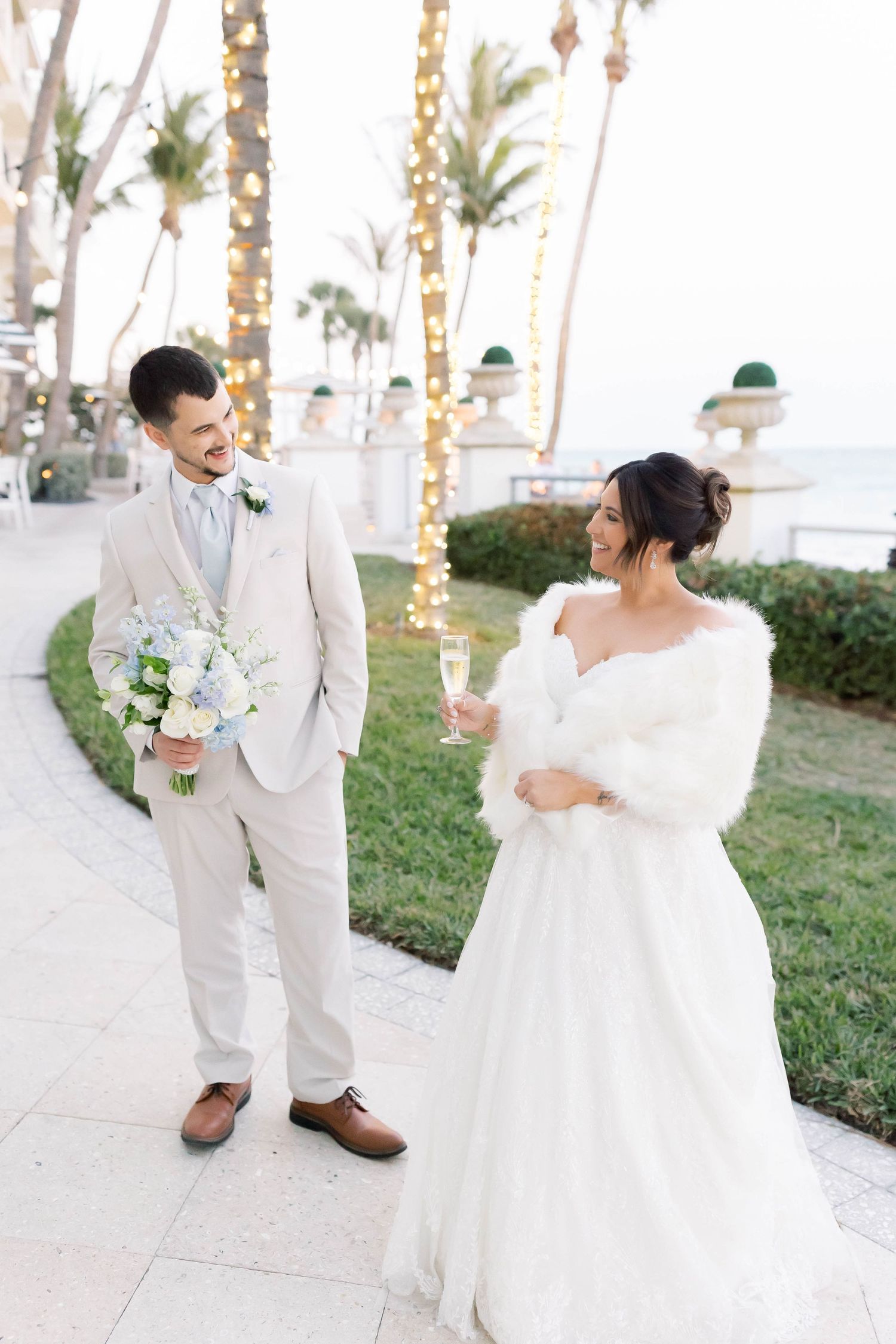 Smiling bride in fur wrap holds champagne as groom carries bouquet near palm trees with string lights.