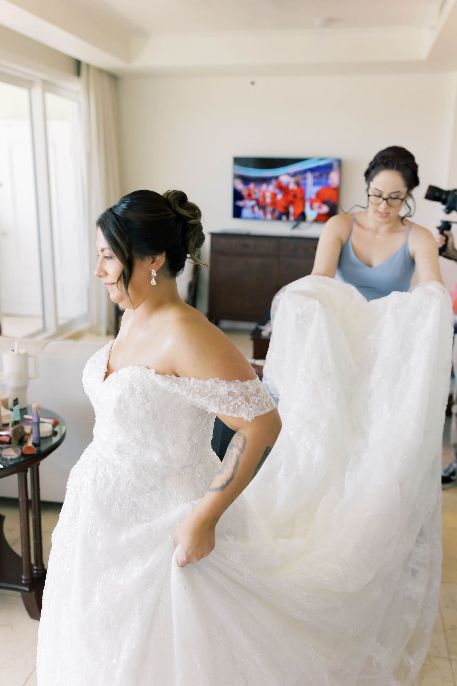 Bride in off-shoulder lace wedding gown being helped with train by bridesmaid in blue dress in hotel room.