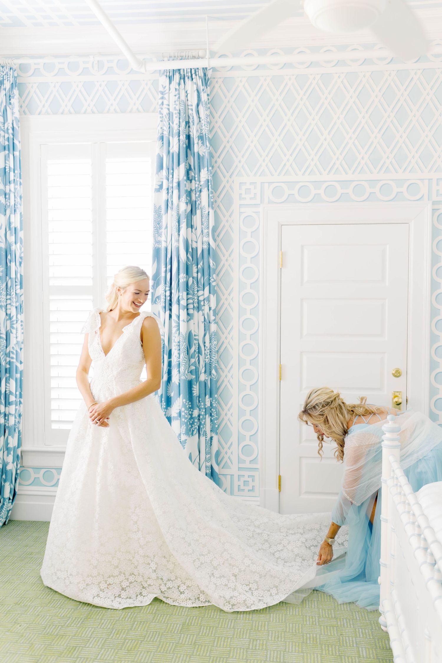 Bride in lace wedding gown laughing in blue wallpapered room while bridesmaid arranges her train.